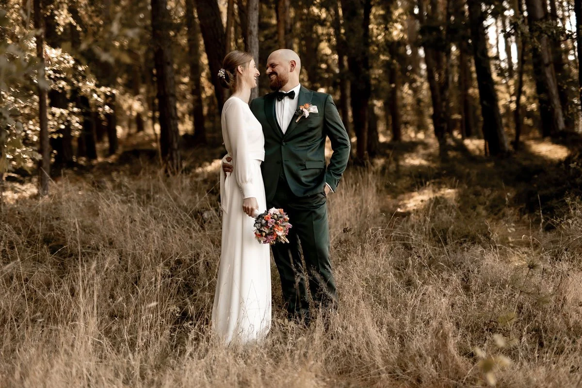 Ein Brautpaar steht in einem Wald, die Braut trägt ein langes weißes Kleid und hält einen Blumenstrauß, der Bräutigam trägt einen schwarzen Anzug mit Fliege.