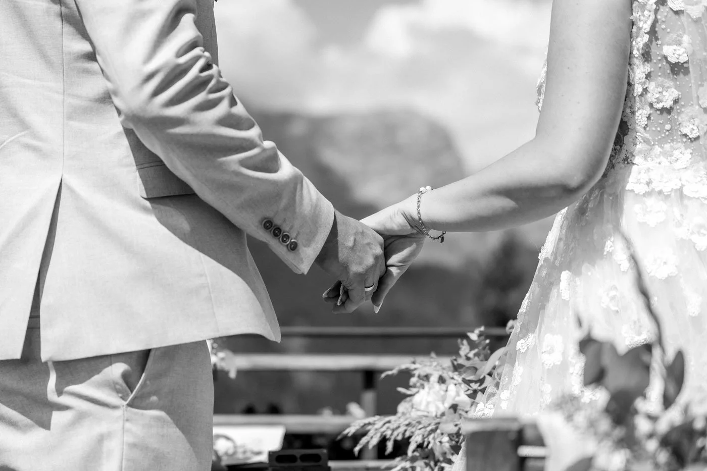Ein Blick auf die Hände eines Paares während einer Hochzeitszeremonie, sie halten sich bei einer Trauung in einem Freiluftumfeld. Das Brautpaar trägt formelle Kleidung, die Braut hat eine Schmuckarmband und ein Kleid mit embossierten Blumen.