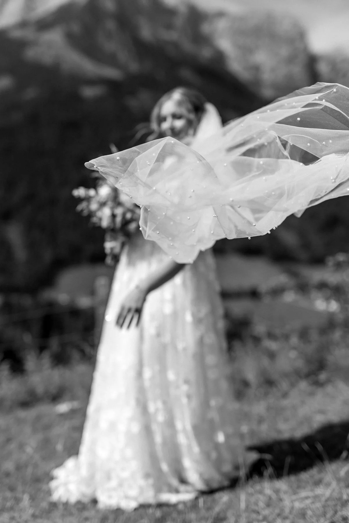 Schwarz-weiß-Foto einer Puppe in einem langen Kleid, die eine große Tüll- oder Organza-Schleife trägt, im Hintergrund eine unscharfe Naturkulisse.
