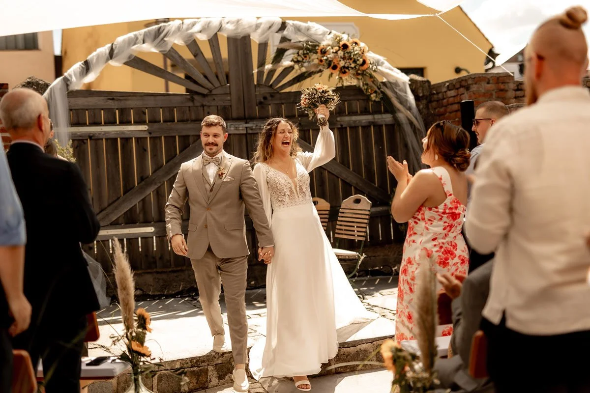 Braut und Bräutigam bei ihrer Hochzeitszeremonie im Freien, umgeben von Freunden und Familie, mit einem Holzportal und Blumen. Die Braut hält den Brautstrauß hoch, alle feiern und applaudieren.