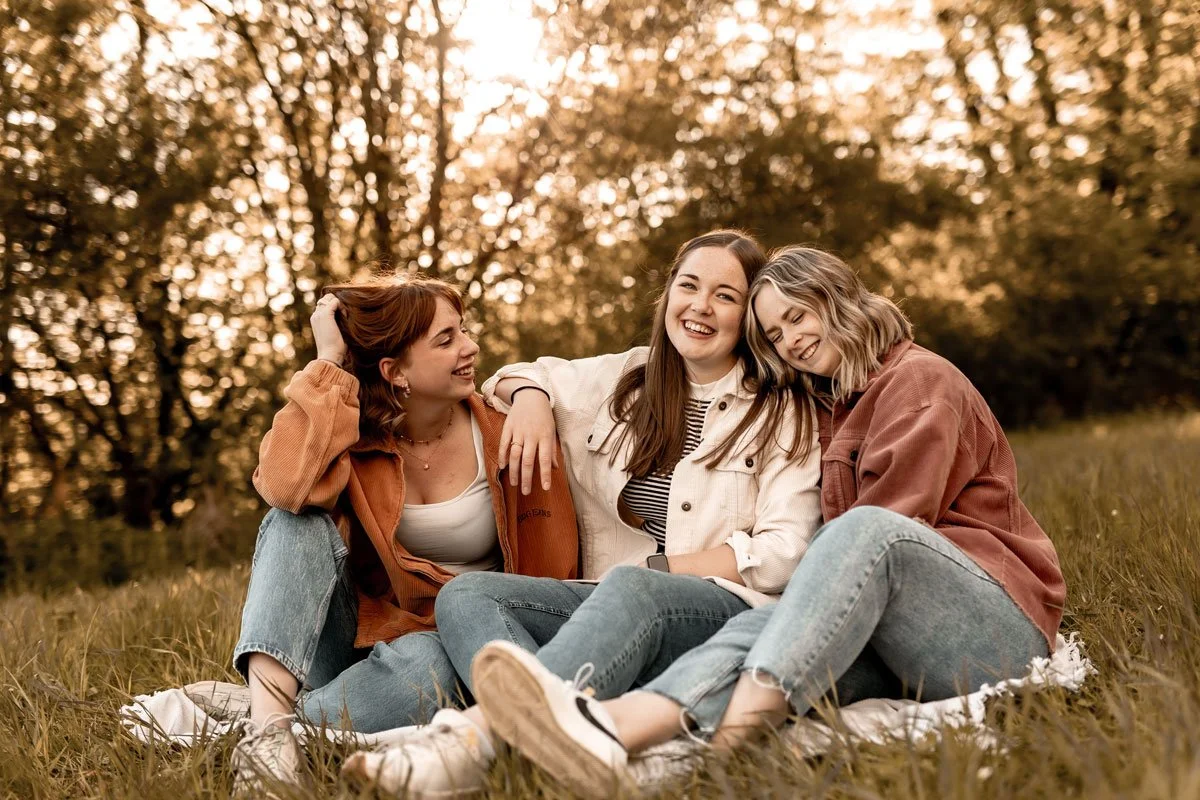 Drei junge Frauen sitzen lachend im Freien auf einer Decke im Gras, umgeben von Bäumen, bei Sonnenuntergang.