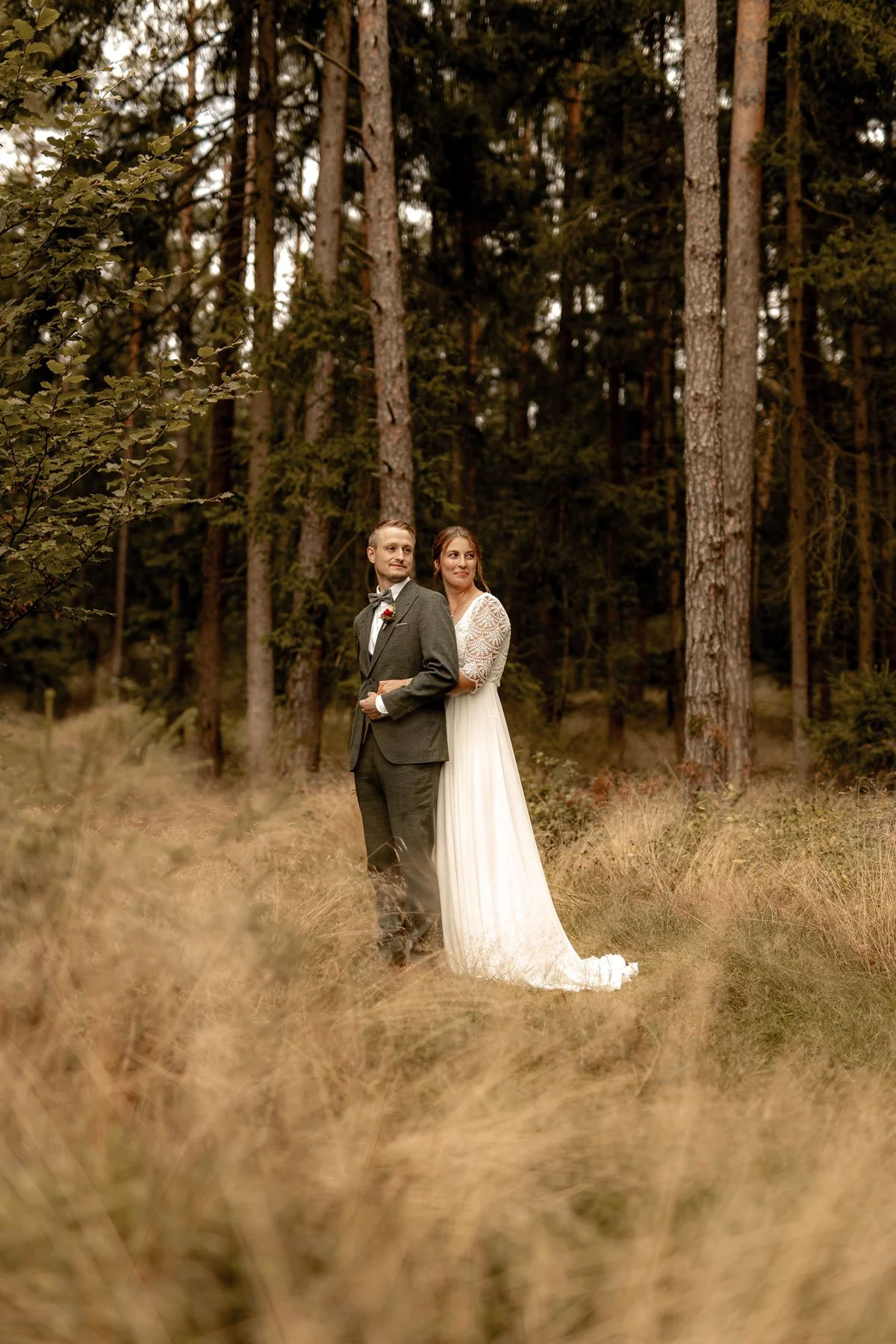 Ein Hochzeitsbild von einem Paar, das in einem Wald steht. Der Bräutigam trägt einen grauen Anzug mit Fliege, die Braut trägt ein langes weißes Hochzeitskleid mit Spitzenärmeln. Beide schauen in verschiedene Richtungen.