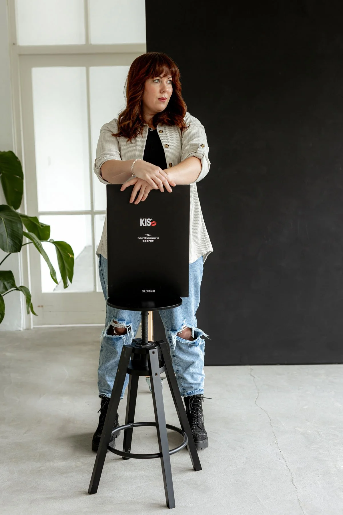 Junge Frau mit roten Haaren in Jeans und Jacke lehnt an Scheinwerfer in einem Raum mit großen Fenstern und schwarzen Hintergrund.