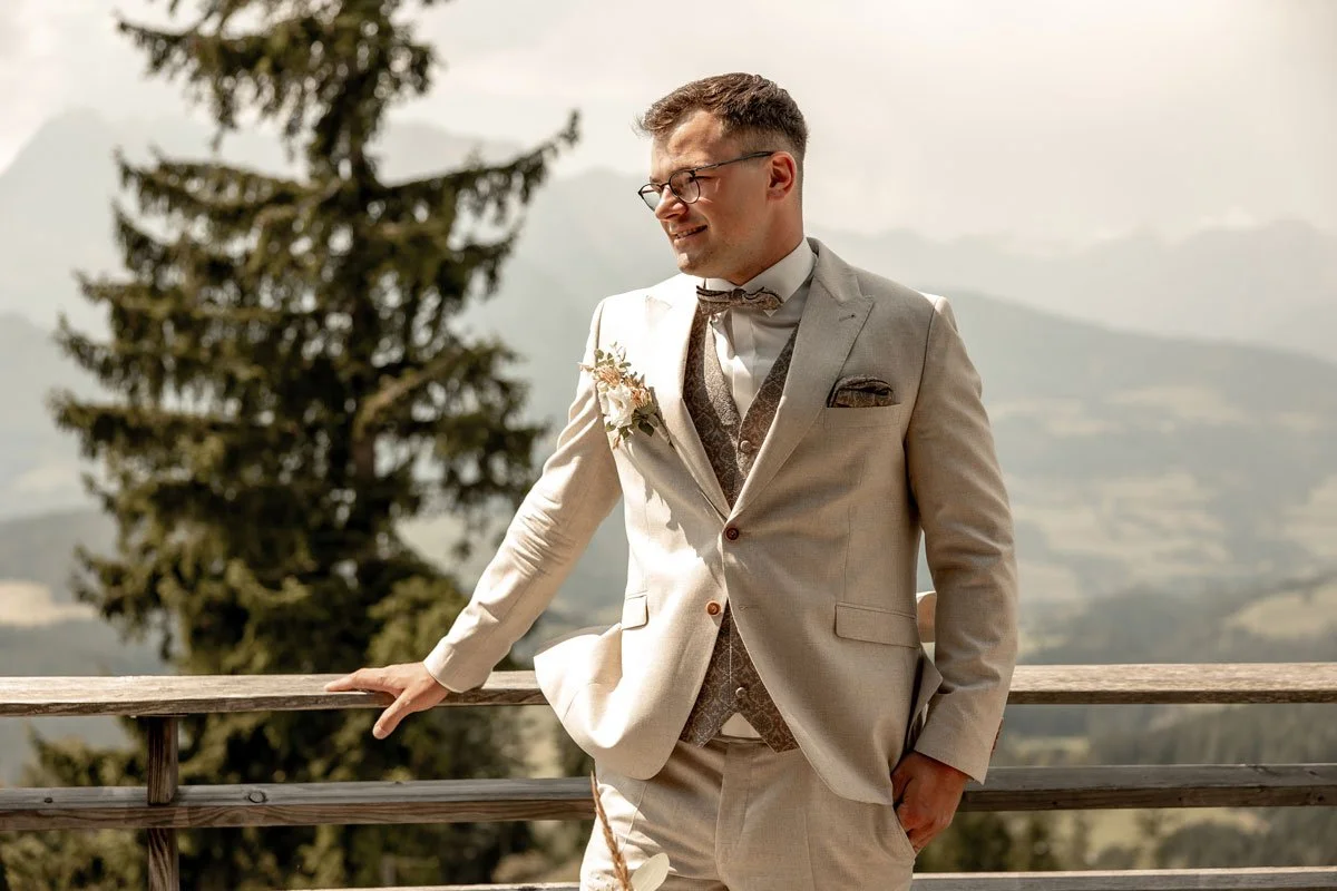 Ein Mann in Hochzeitskleidung, der auf einer Holzbrüstung steht, im Freien mit Bergen und Bäumen im Hintergrund.