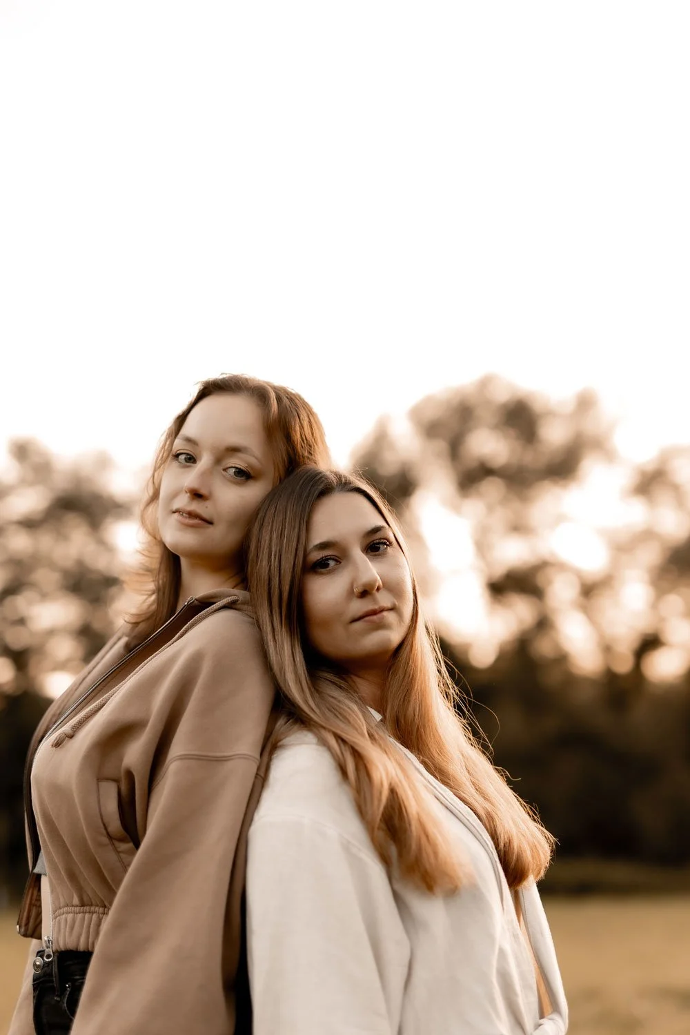 Zwei junge Frauen stehen eng beieinander im Freien bei Sonnenuntergang, vor einem sommerlichen Baumhintergrund.