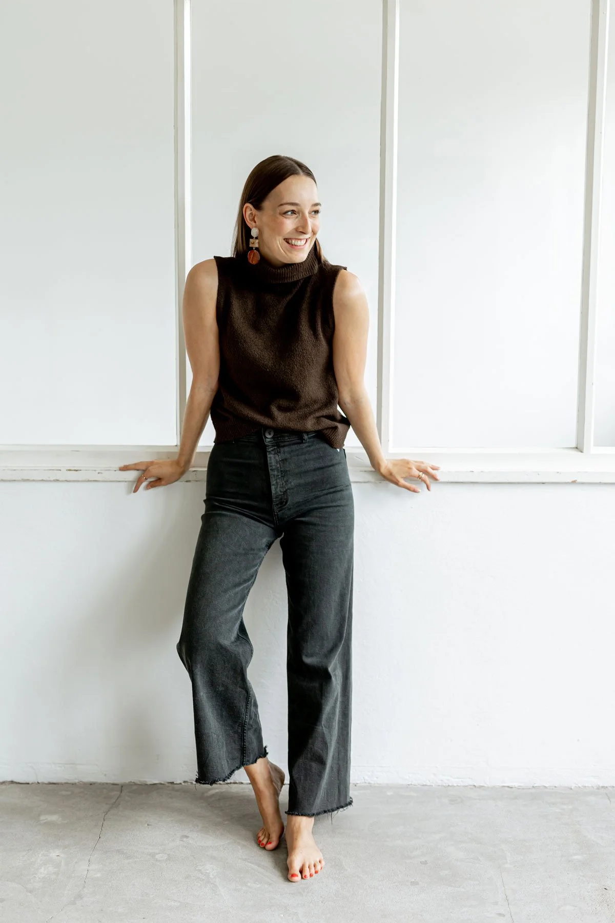 Eine lachende Frau mit dunklen Haaren, trägt ein braunes ärmelloses Oberteil und schwarze Jeans, steht barfuß an einem weißen Fensterrahmen in einem Raum mit hellem Boden.