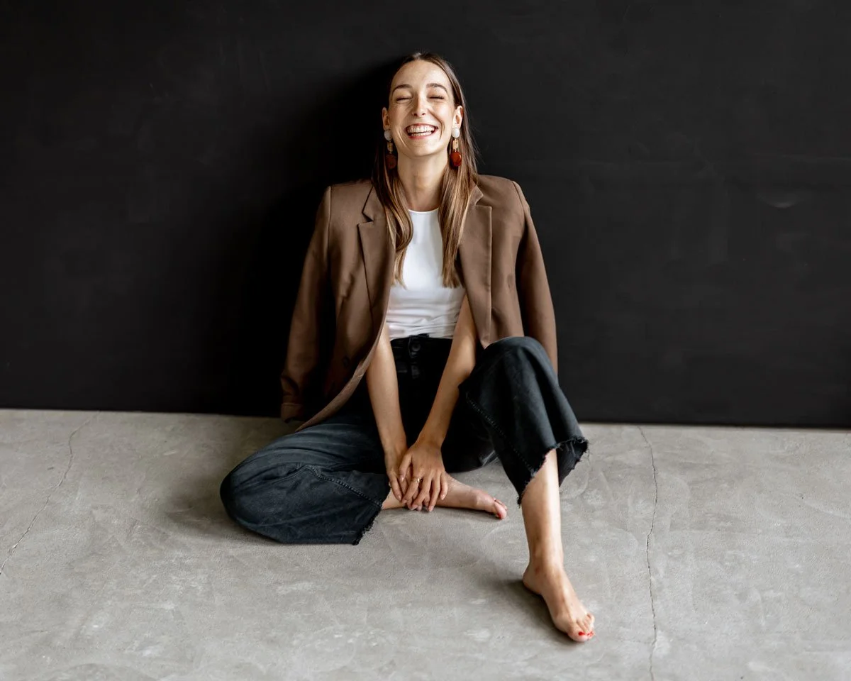 Lächelnde Frau sitzt nackt und leger auf dem Boden vor einer schwarzen Wand.