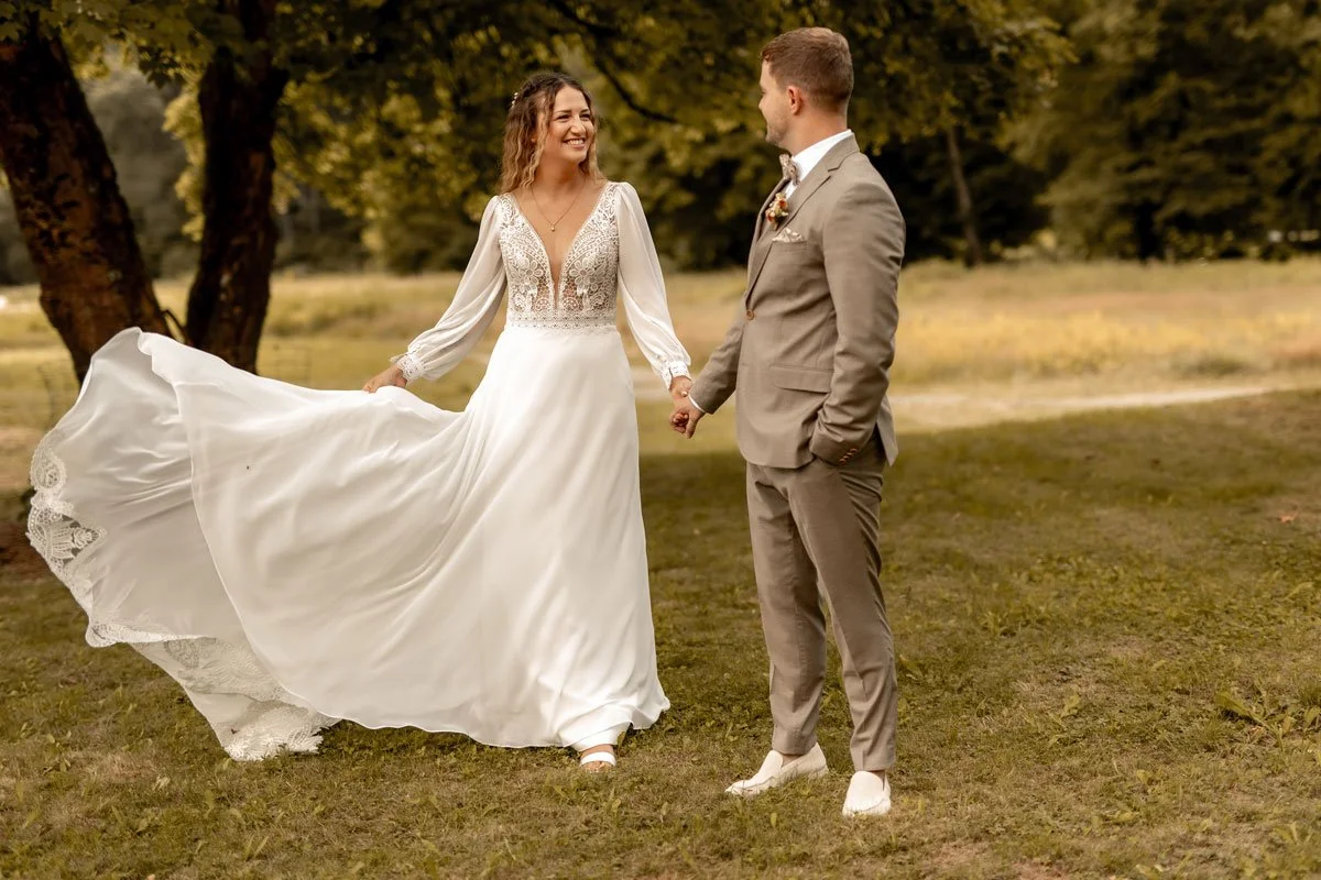 Ein Brautpaar hält Händchen im Grünen. Die Braut trägt ein weißes, langes Hochzeitskleid mit Spitze, der Bräutigam einen beigen Anzug. Es ist ein sonniger Tag im Park.