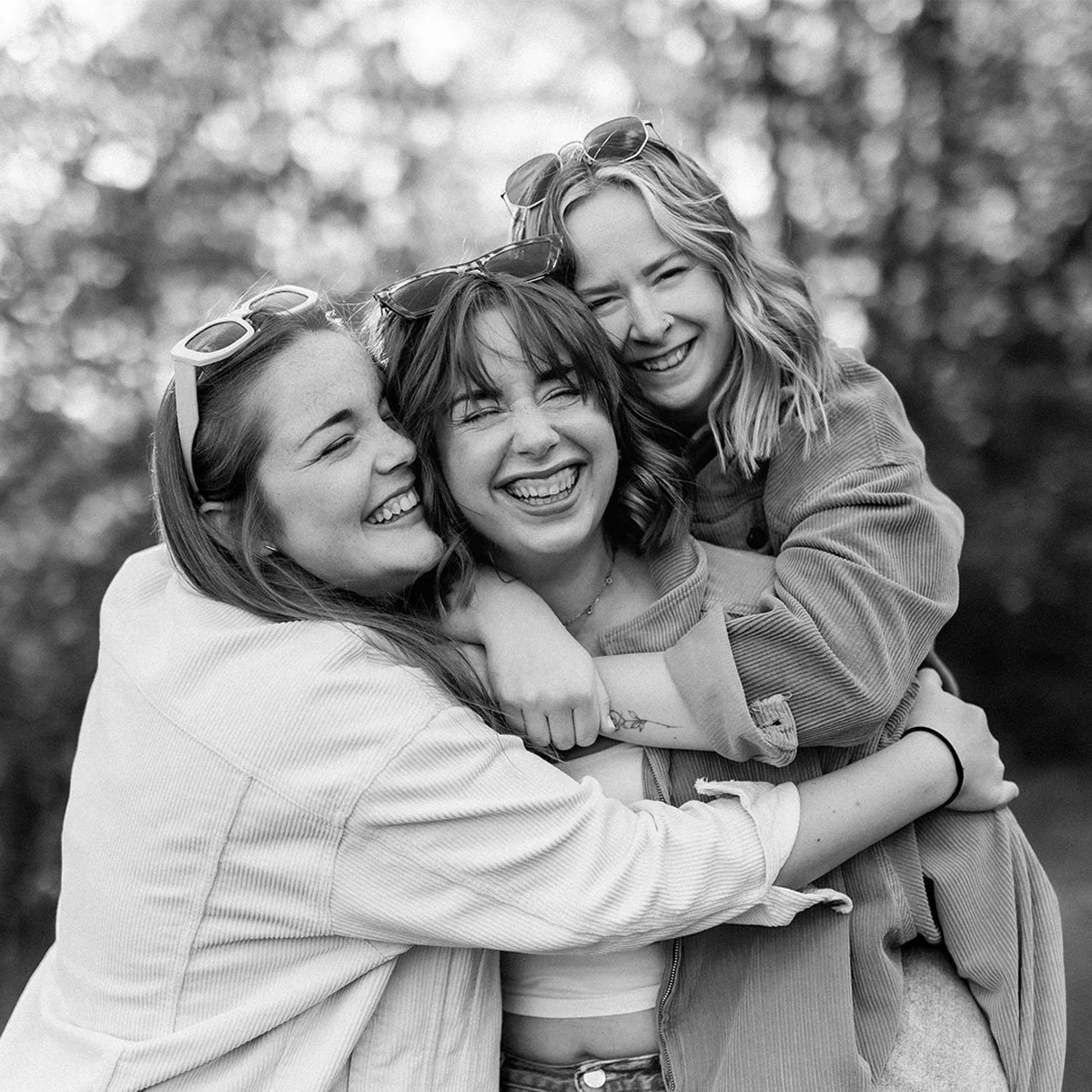 Drei lachende Frauen umarmen sich draußen in der Natur, alle tragen Sonnenbrillen auf dem Kopf.