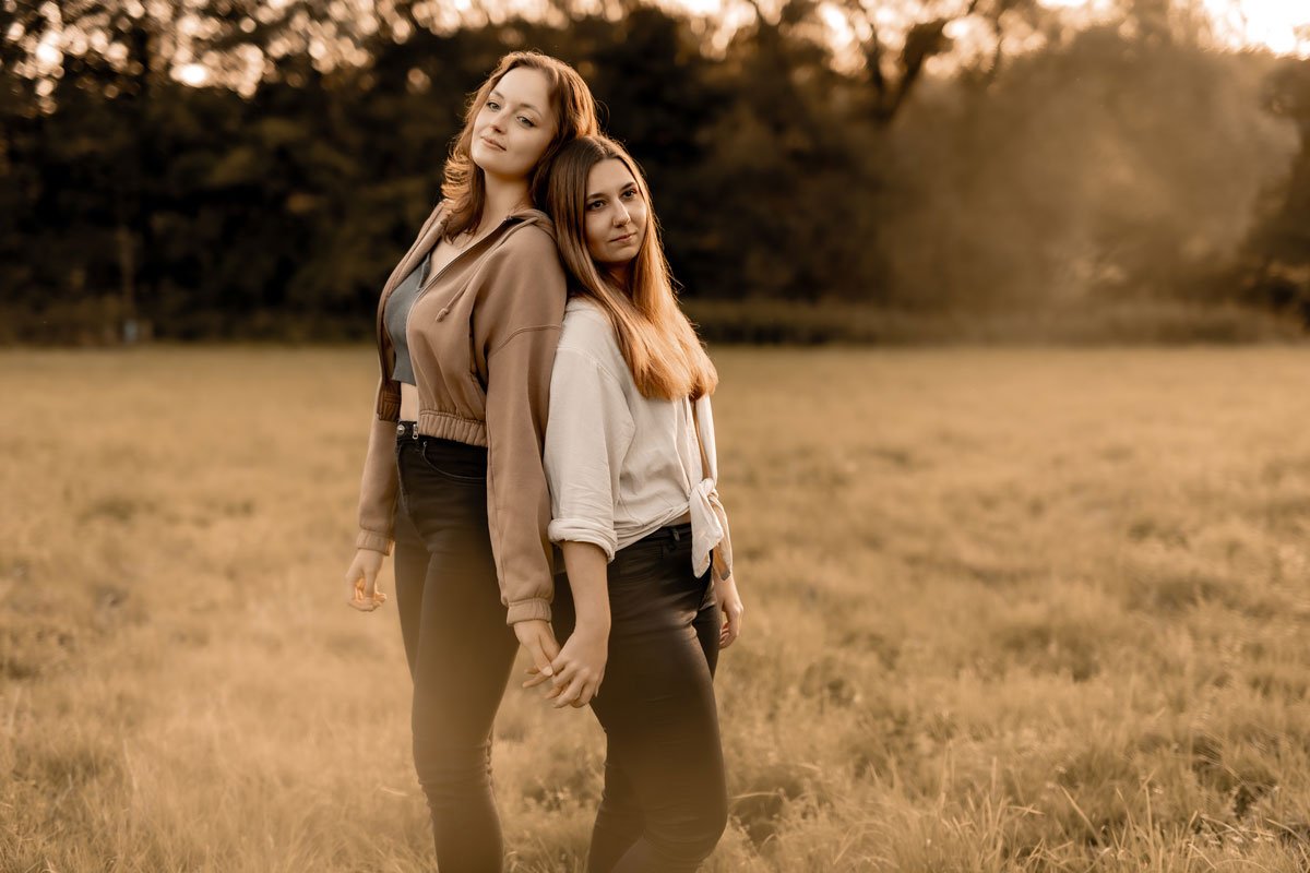 Zwei junge Frauen stehen im Sonnenuntergang in einem offenen Feld, halten sich an den Händen und schauen in die Kamera.