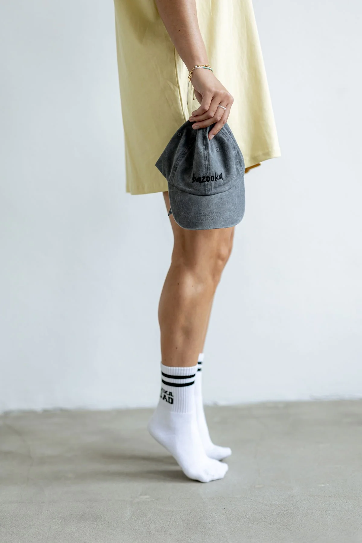Person hält eine graue Baseballkappe mit der Aufschrift 'bazooka', trägt weiße Sportsocken mit schwarzen Streifen und einem Text auf der Seite, trägt einen gelben Kleid oder Rock, steht auf einem grauen Boden gegen eine weiße Wand.