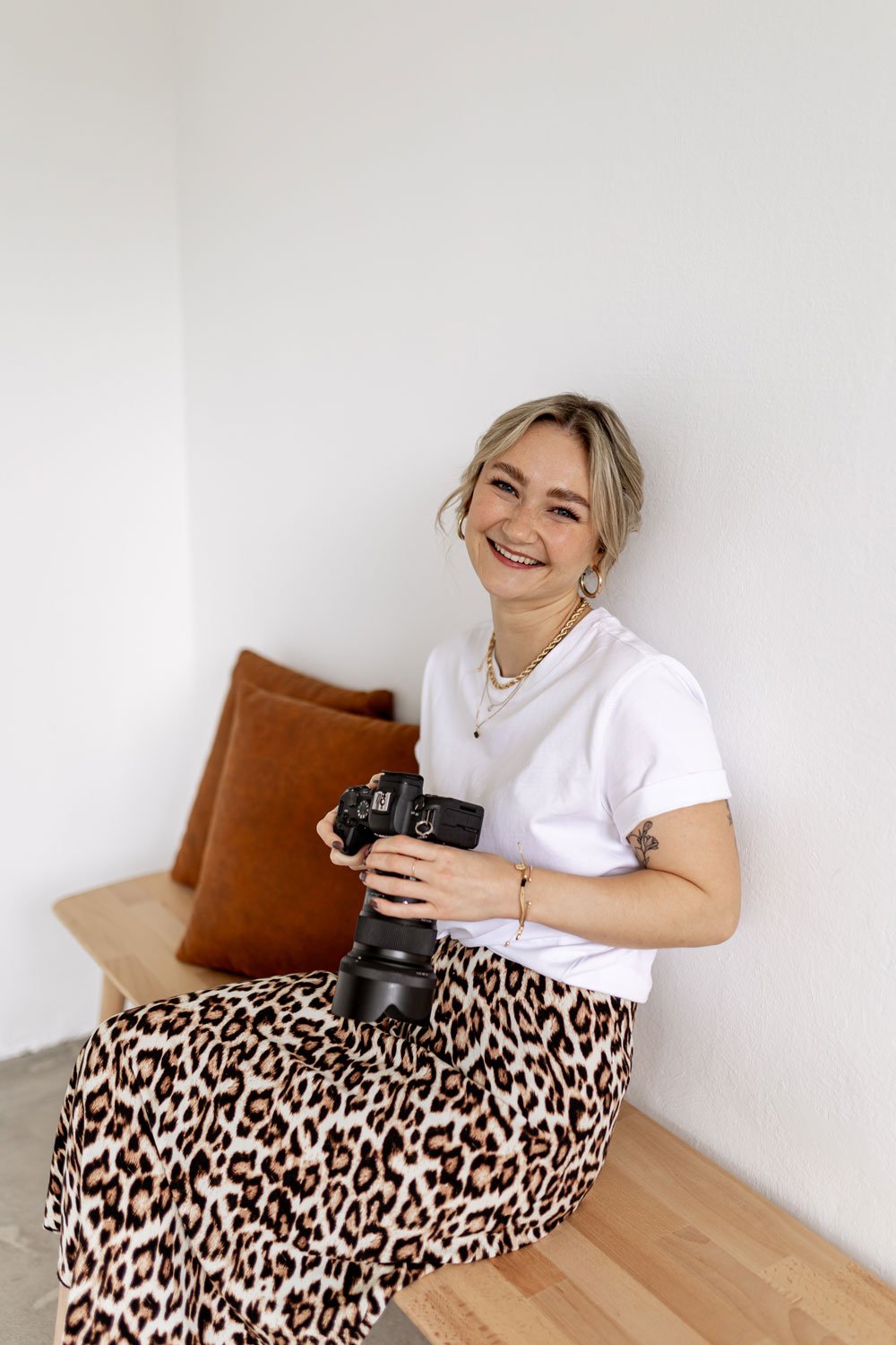 Eine lächelnde Frau mit kurzen blondem Haar, die in einem weißen T-Shirt sitzt, ein Kamera in den Händen hält, vor einer weißen Wand und auf einem Holzbank mit braunen Kissen.