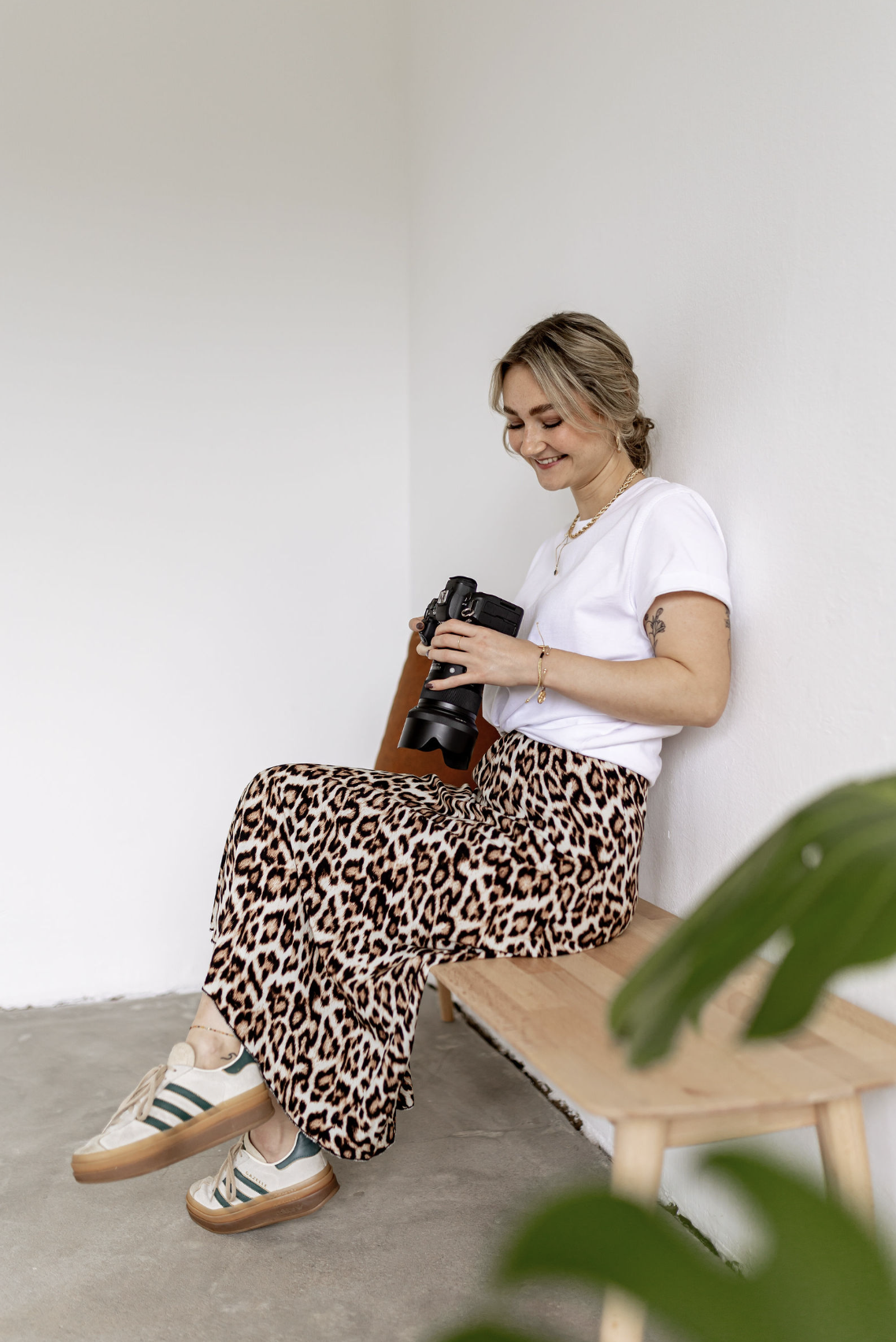 Junge Frau sitzt auf einer Bank, hält eine Kamera in der Hand, trägt ein weißes T-Shirt, eine Leoparden-Jeans und weiße Turnschuhe mit grünen Streifen, lächelt.
