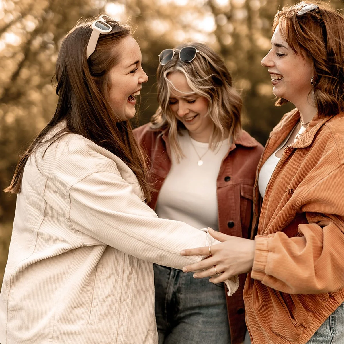 Drei lachende Frauen, die sich im Freien umarmen, in einem warmen Farbton mit Herbstbäumen im Hintergrund.