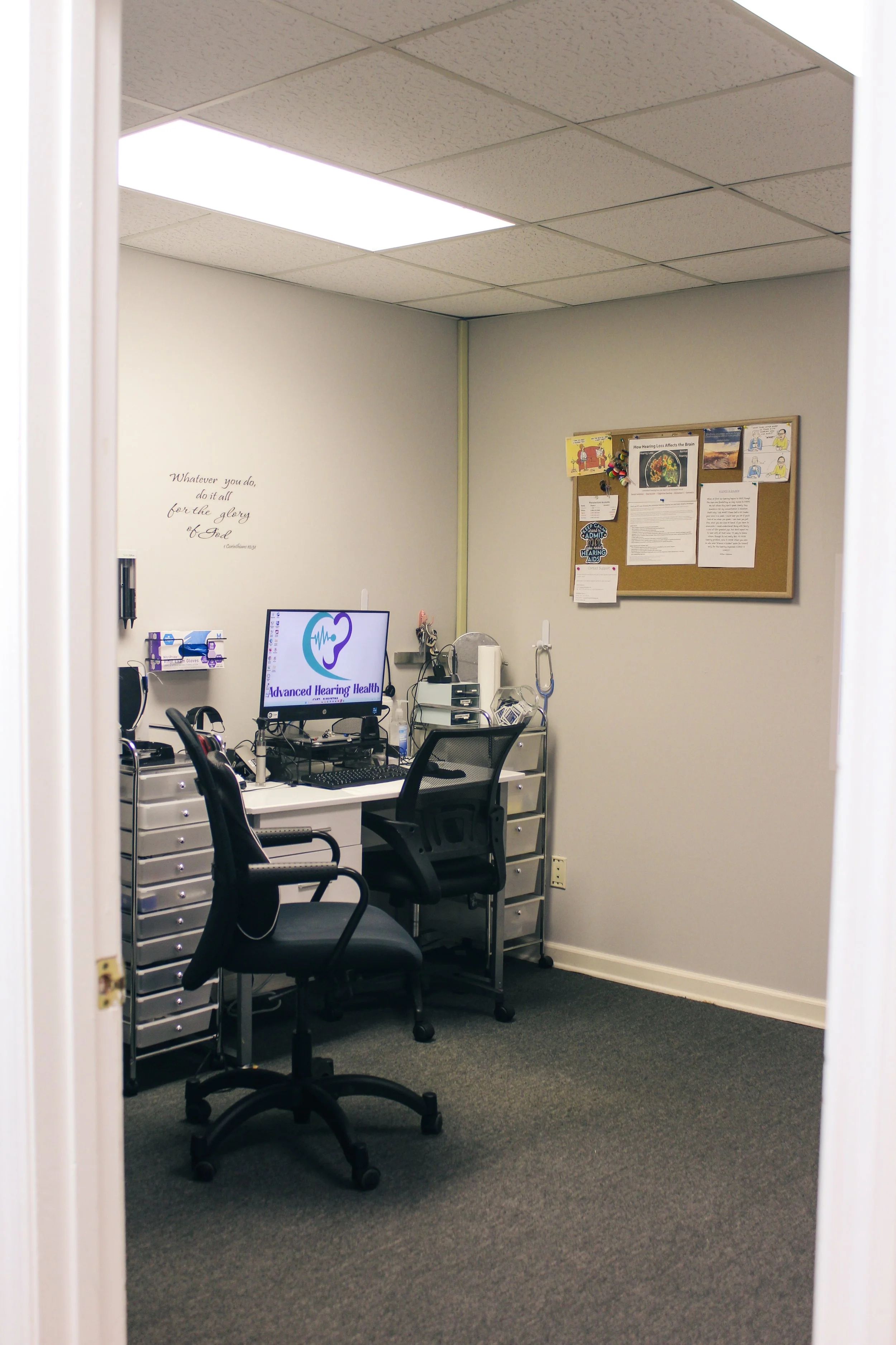 A small office room with two black office chairs, a white desk with computer equipment, and a bulletin board on the wall.