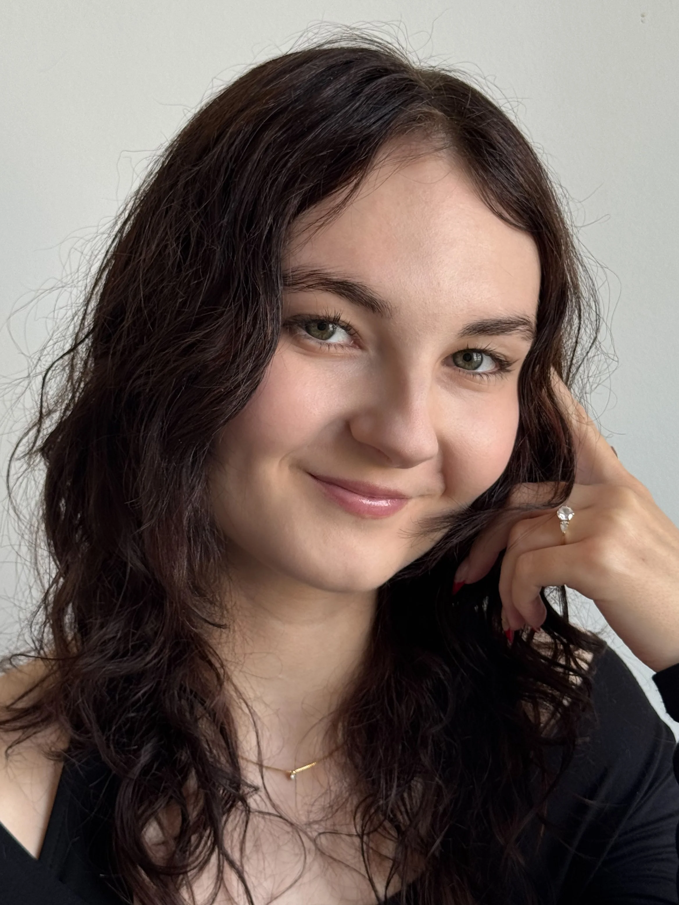 Photo d'une jeune femme aux cheveux bruns ondulés, souriante, portant un collier et une bague, assise contre un fond blanc.