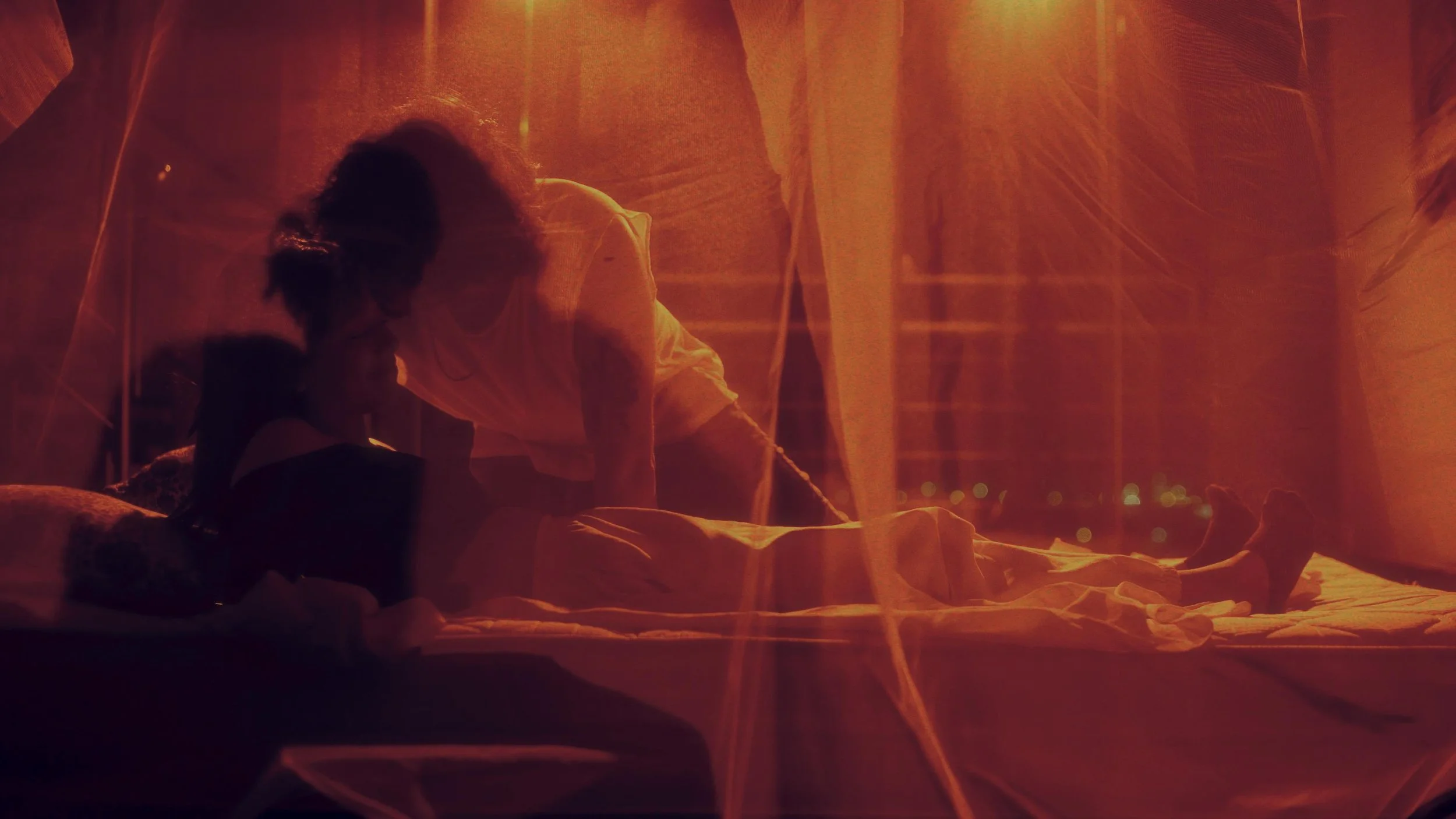 Deux personnes allongées sur un lit, dans une pièce éclairée d'une lumière orange, avec des rideaux et des drapes en arrière-plan, créant une ambiance chaleureuse et intime.