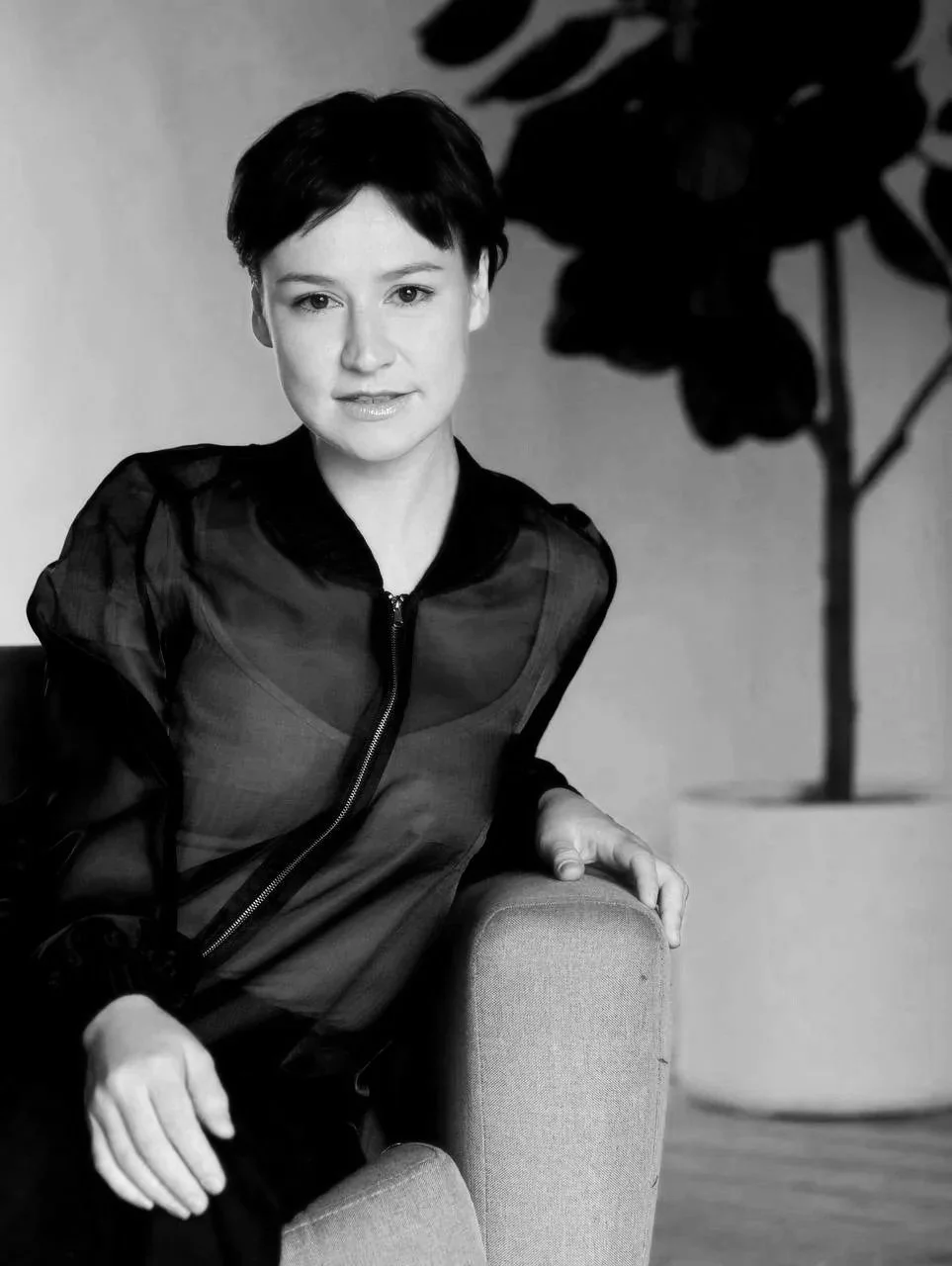 A black-and-white portrait of a woman with short dark hair sitting on a chair, wearing a dark satin-like jacket, with a large potted plant in the background.