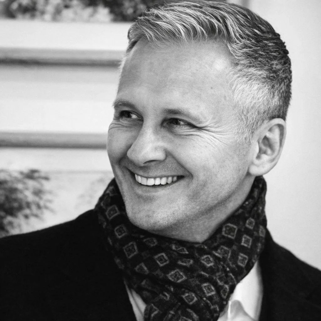 Black and white photo of a smiling man with short, gray hair, wearing a patterned scarf and a dark jacket.