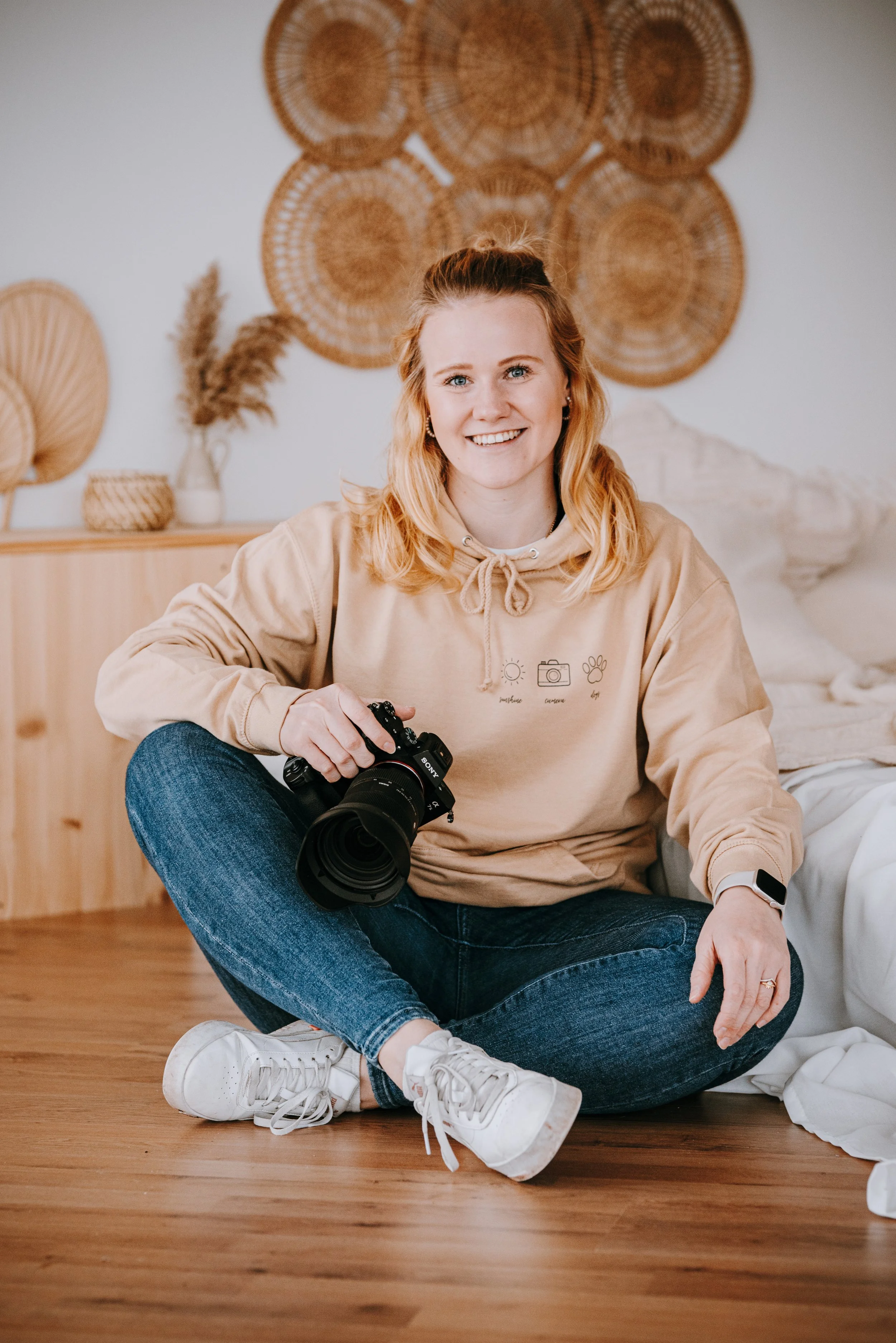 Junge Frau sitzt auf dem Boden, hält eine Kamera in der Hand und lächelt, mit decorativen Wandbildern und Vasen im Hintergrund.