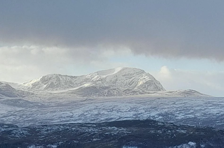 A snow covered Ben Hope under a cloudy sky, with a dark, rugged terrain in the foreground.