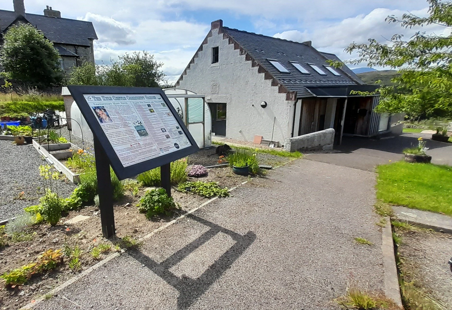 The Kyle Centre - a white house with a pitched roof and skylights, surrounded by a garden with potted plants and flowers. There is a signboard with text and photos in the foreground, and a pathway leading to the house under a partly cloudy sky.