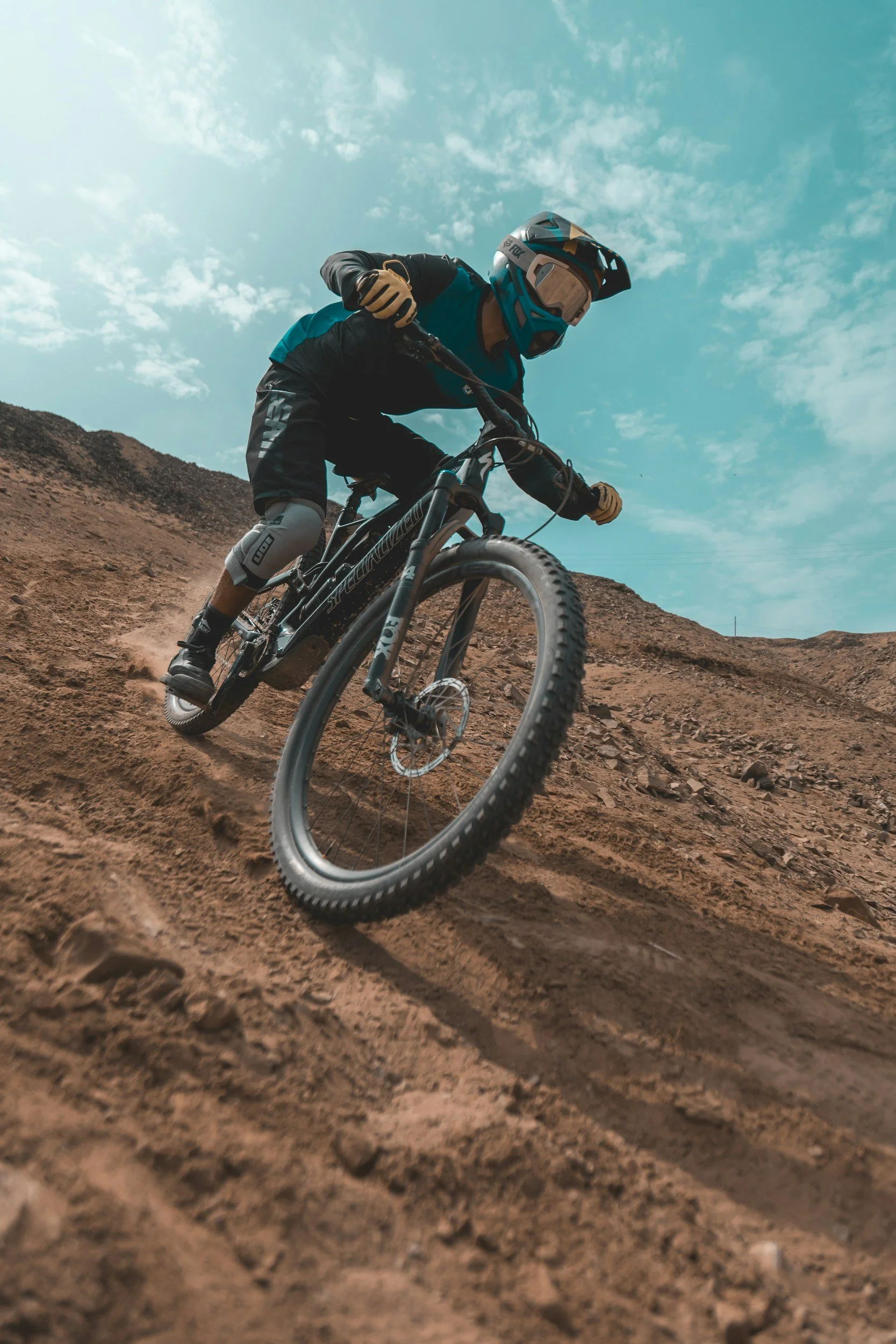A mountain biker wearing a helmet, protective gear, and a blue and black outfit rides down a dirt trail on a mountain under a partly cloudy sky.