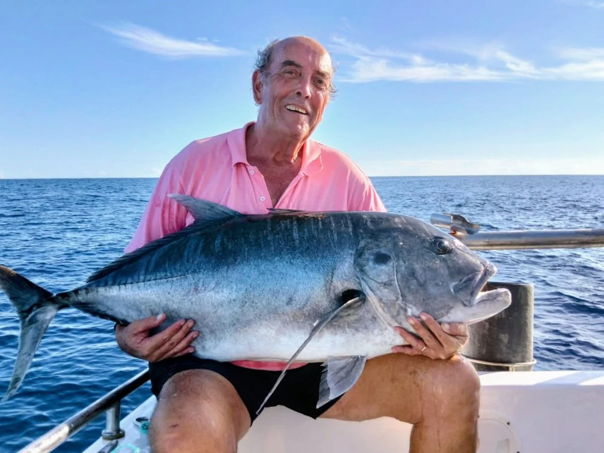 An elderly man smiling while sitting on a boat holding a large fish he caught, with the ocean and a blue sky in the background.