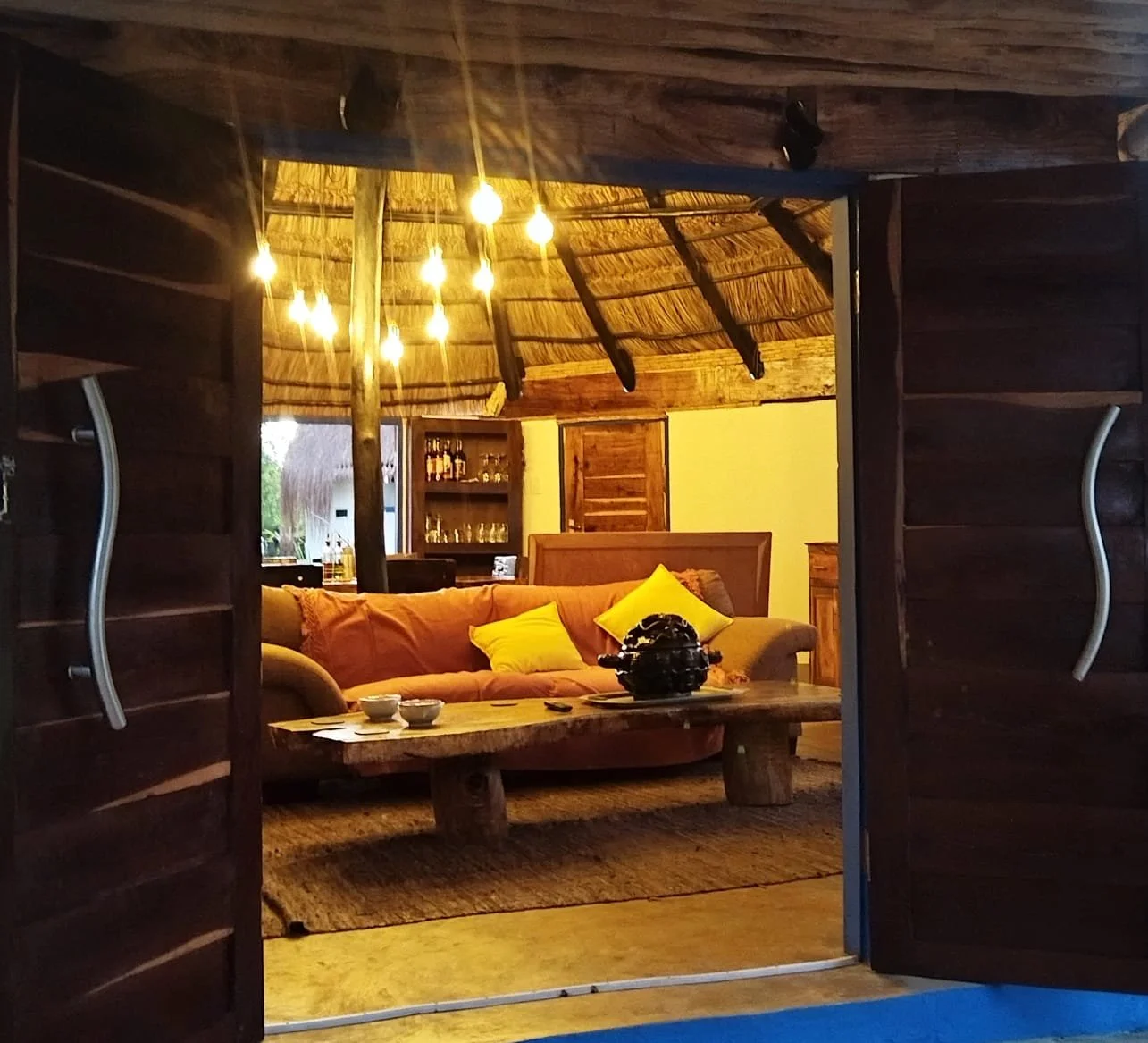 A cozy living room seen through a door with curved metal handles, featuring a pink sofa with yellow pillows, a rustic wooden coffee table with items, and a thatched ceiling with hanging lights.