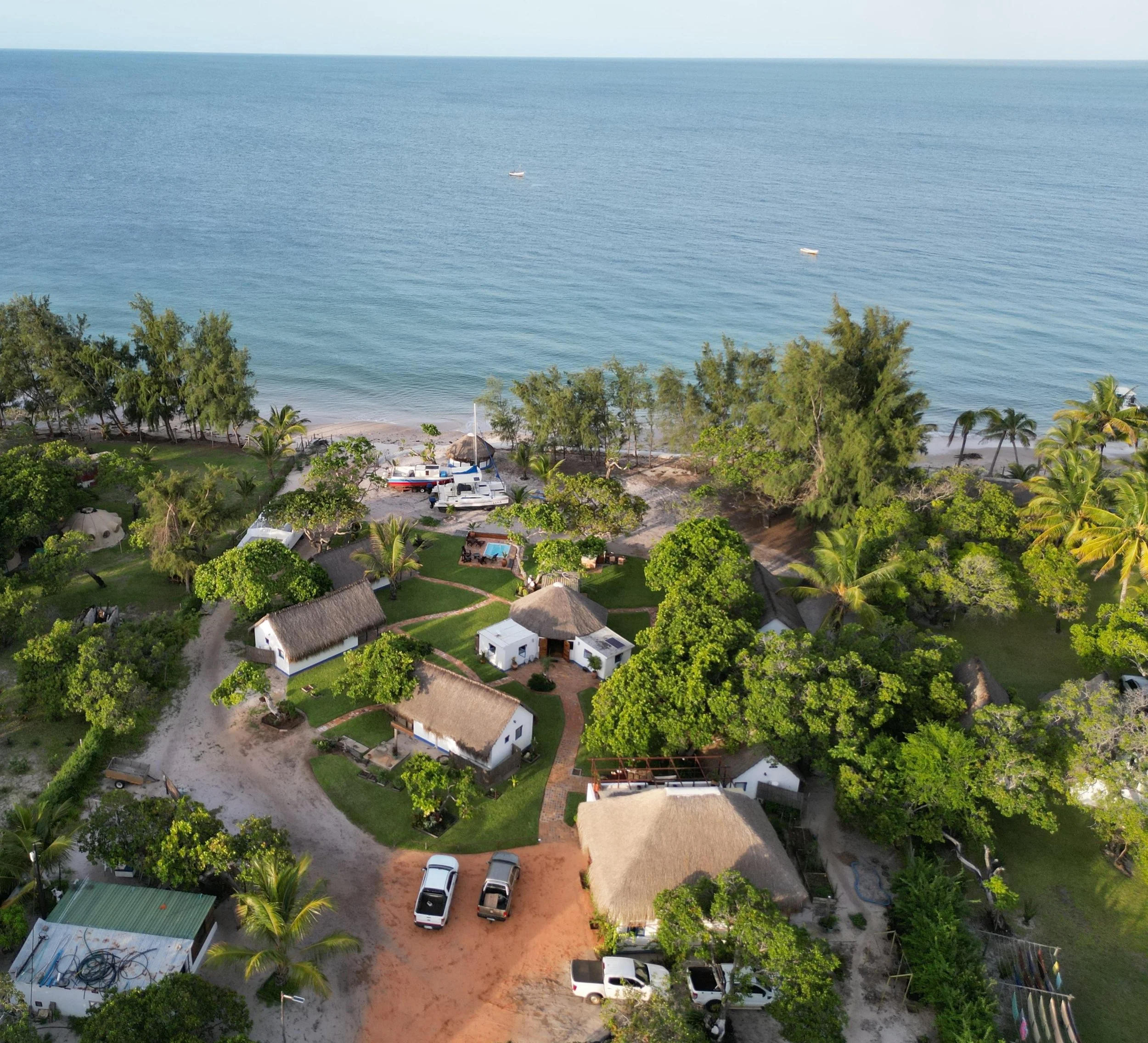 Aerial view of a beachside resort with green trees, sandy pathways, parked vehicles, and a view of the ocean with two boats in the water.