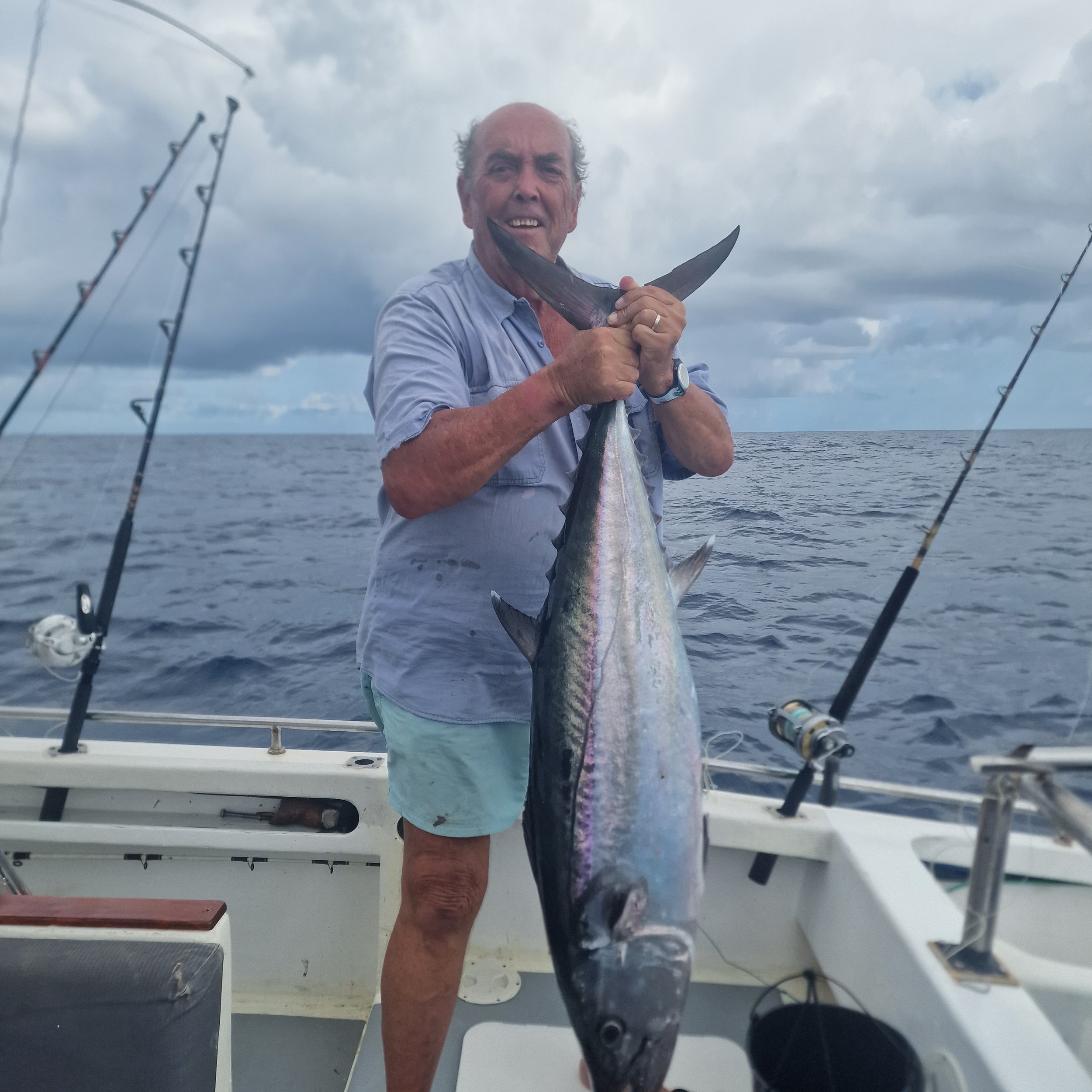 A man on a boat holding a large fish, with fishing rods and cloudy sky in the background.