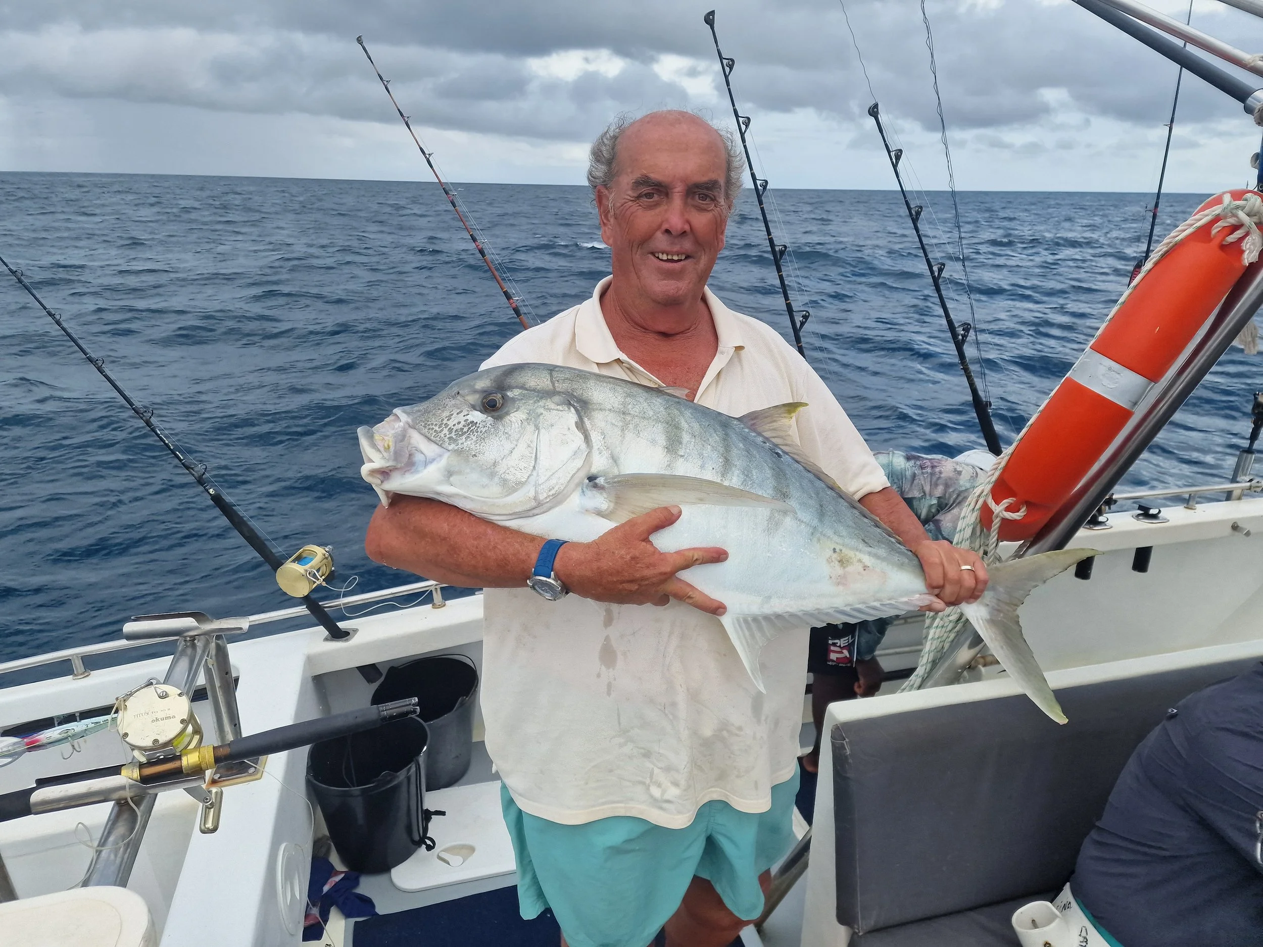 An older man holding a large fish on a boat at sea with fishing rods in the background, under cloudy skies.