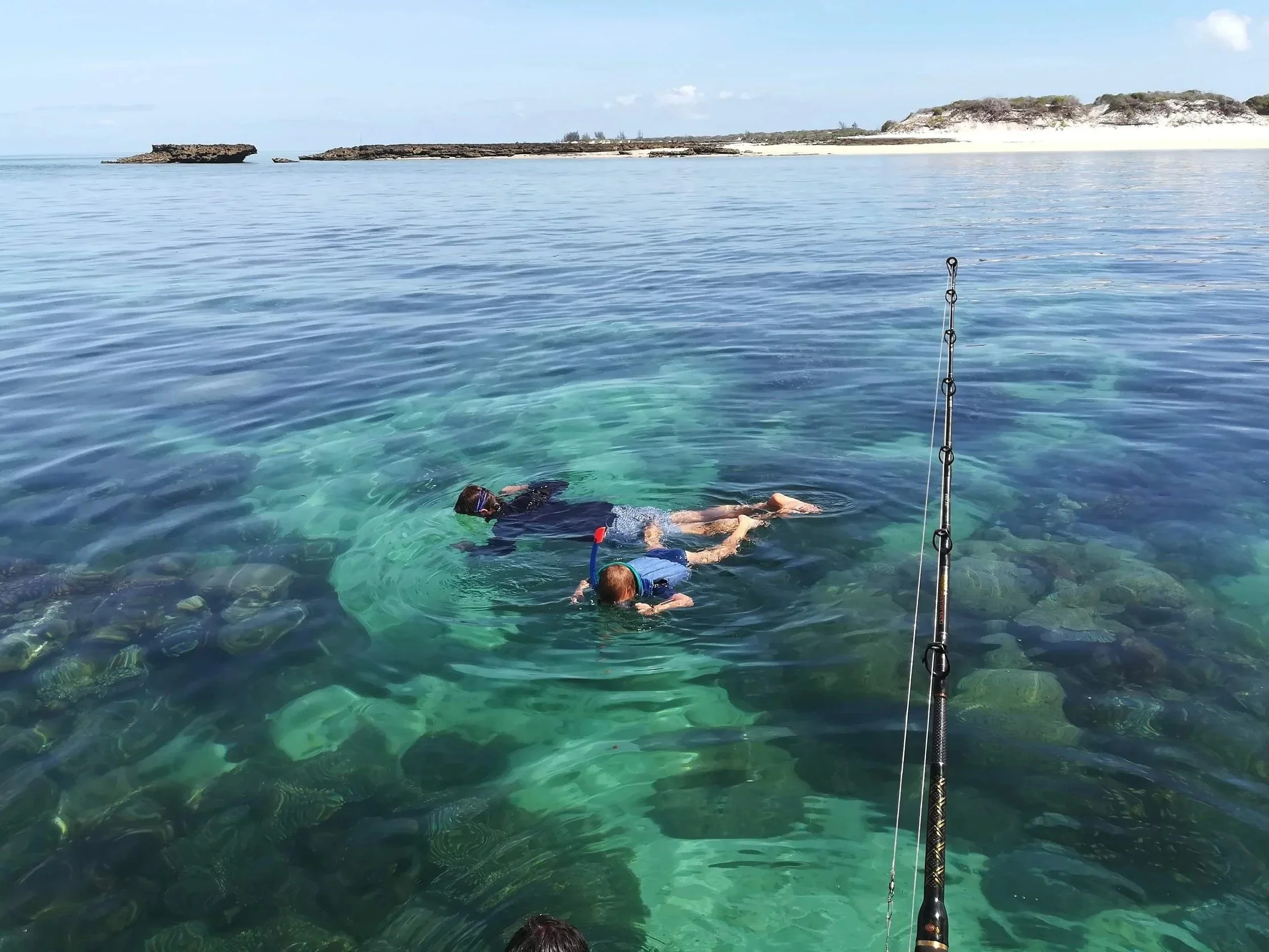 People snorkeling in clear ocean water near rocky coastline with white sandy beach and cliffs in the background.