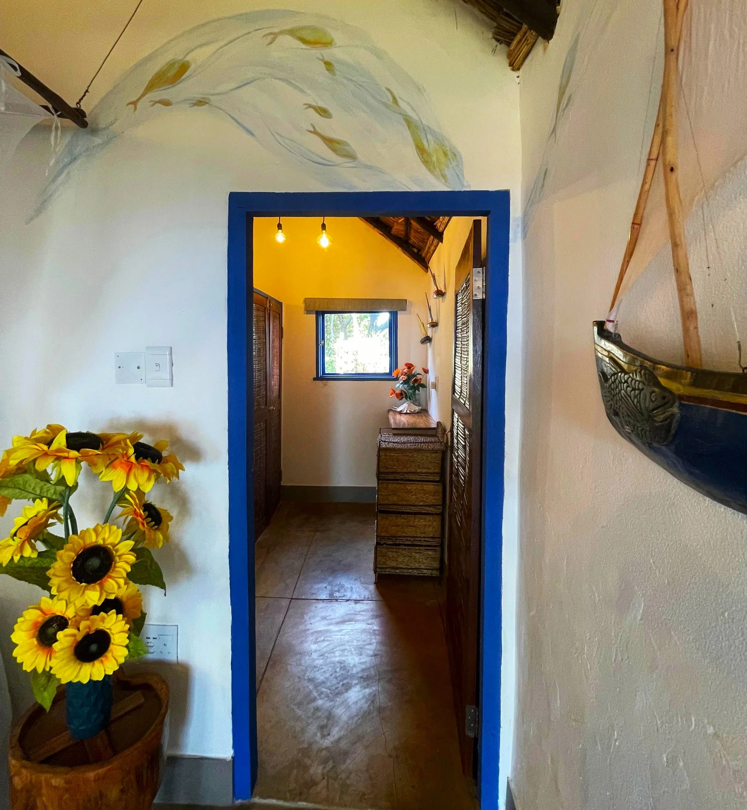View through a doorway into a room with yellow walls, a window with blinds, and decorative vases on the wall. In the foreground on the left, there is a blue vase filled with large, bright yellow flowers. To the right, a wooden or cane bench is visibl