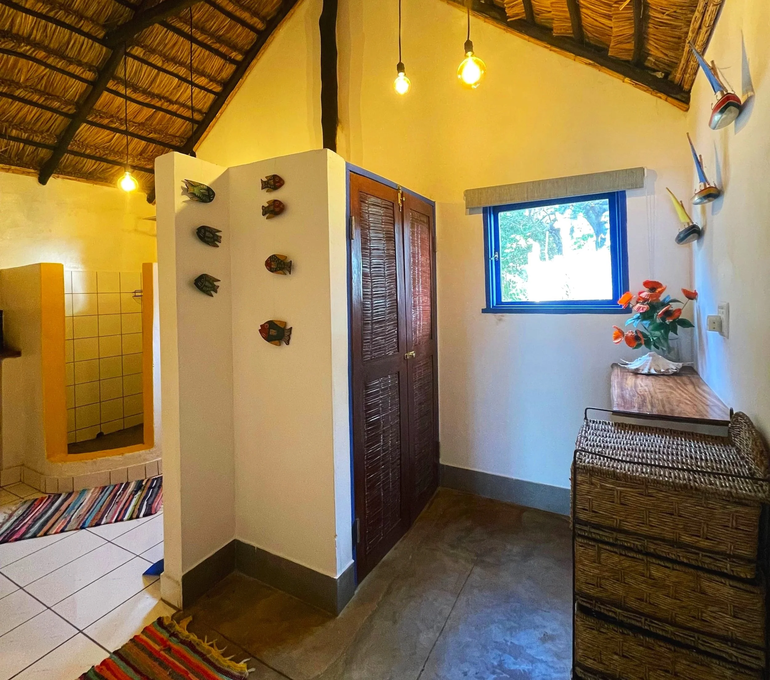 Interior of a room with a thatched roof, yellow walls, hanging light bulbs, and a window revealing greenery outside. There are decorative fish wall hangings, wicker furniture, and colorful rugs.