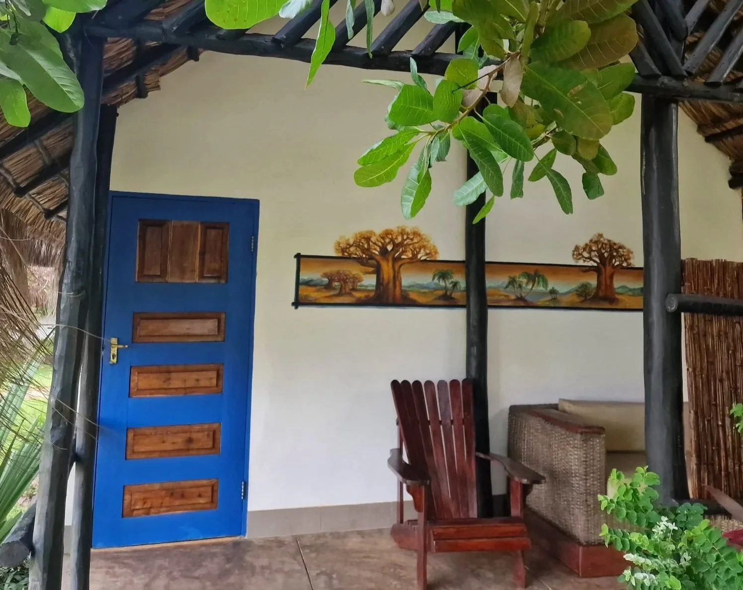 Interior of a rustic room with a blue door, a mural of baobab trees on the wall, a wooden chair, a wicker bench, and green plants, with a thatched roof overhead.