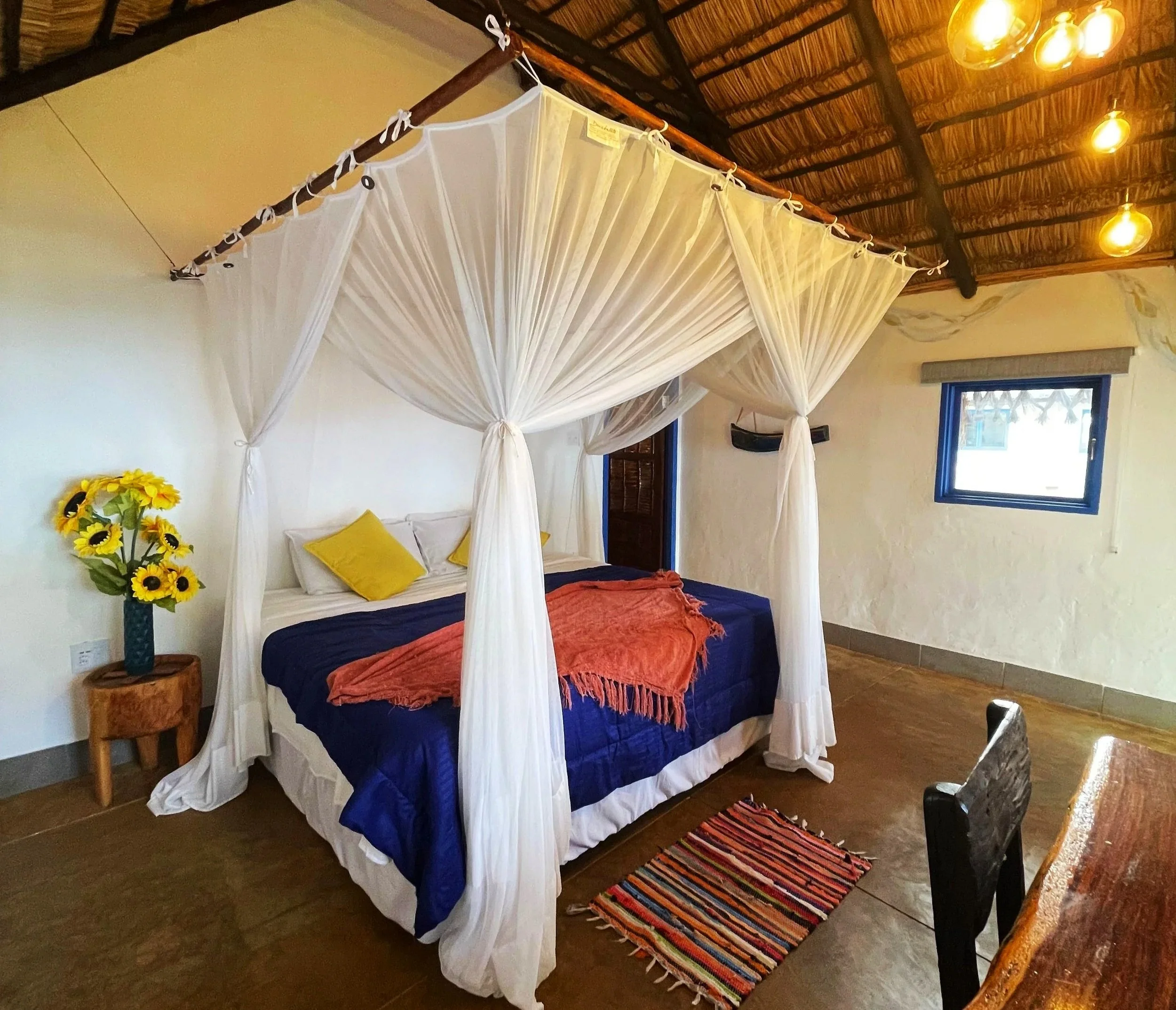 A bedroom featuring a white canopy bed with cream curtains, decorated with yellow pillows, a reddish-orange blanket, and a small bouquet of sunflowers in a vase on a wooden side table. The room has a woven bamboo ceiling with wooden beams, yellow han
