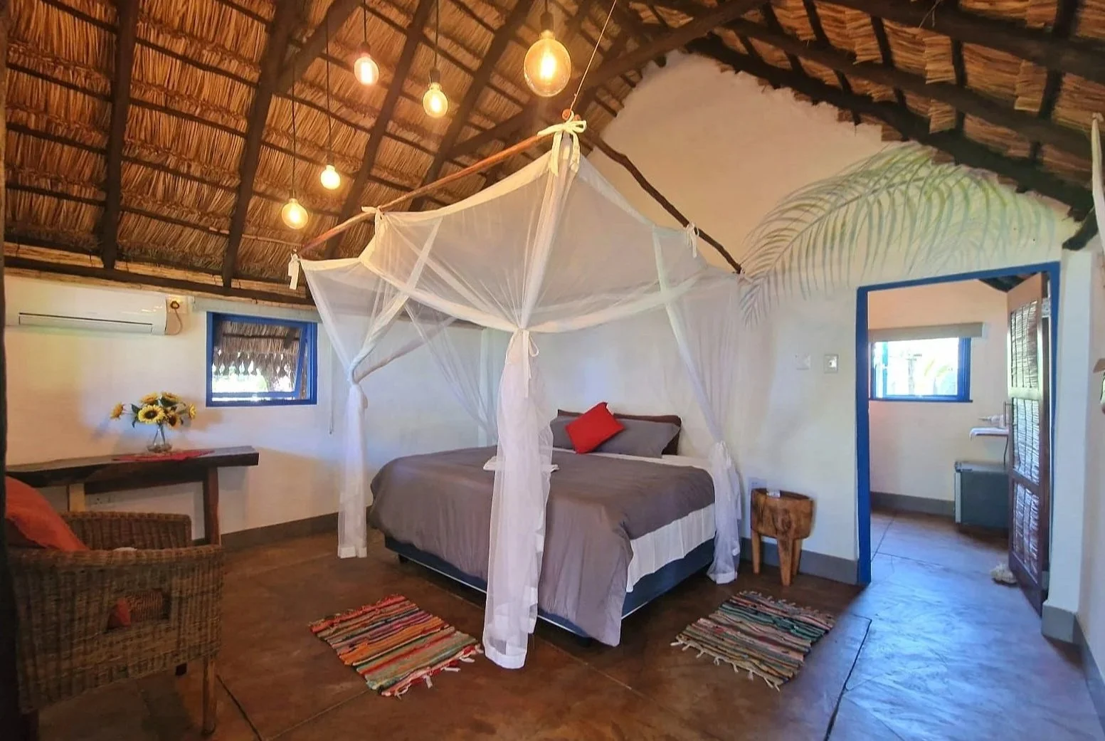 A cozy bedroom with a thatched roof ceiling, a four-poster bed with white mosquito netting and a red pillow, small colorful rugs on a wooden floor, a wicker chair, a wooden table with a sunflower vase, and a view into an adjoining room with a window.