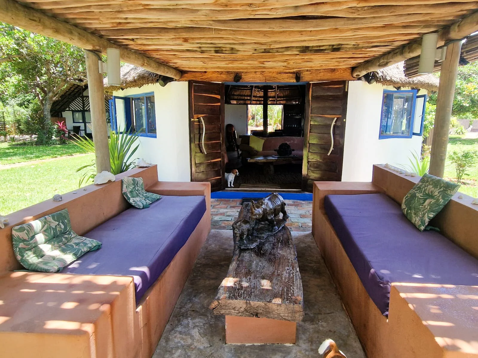 Outdoor living area with two orange sofas with purple cushions and green patterned pillows, a rustic wooden coffee table, open wooden doors revealing a cozy interior with a dog inside, surrounded by lush greenery and a thatched roof.