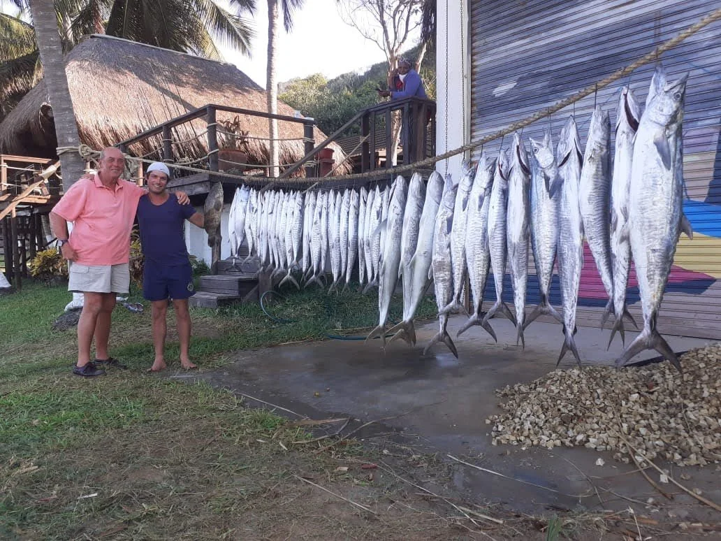 Two men standing in front of a string of hanging fish outdoors. One man is wearing a pink shirt and shorts, the other is wearing a blue shirt and shorts. There is a person on a balcony behind them, and a small hut with a thatched roof is visible in t