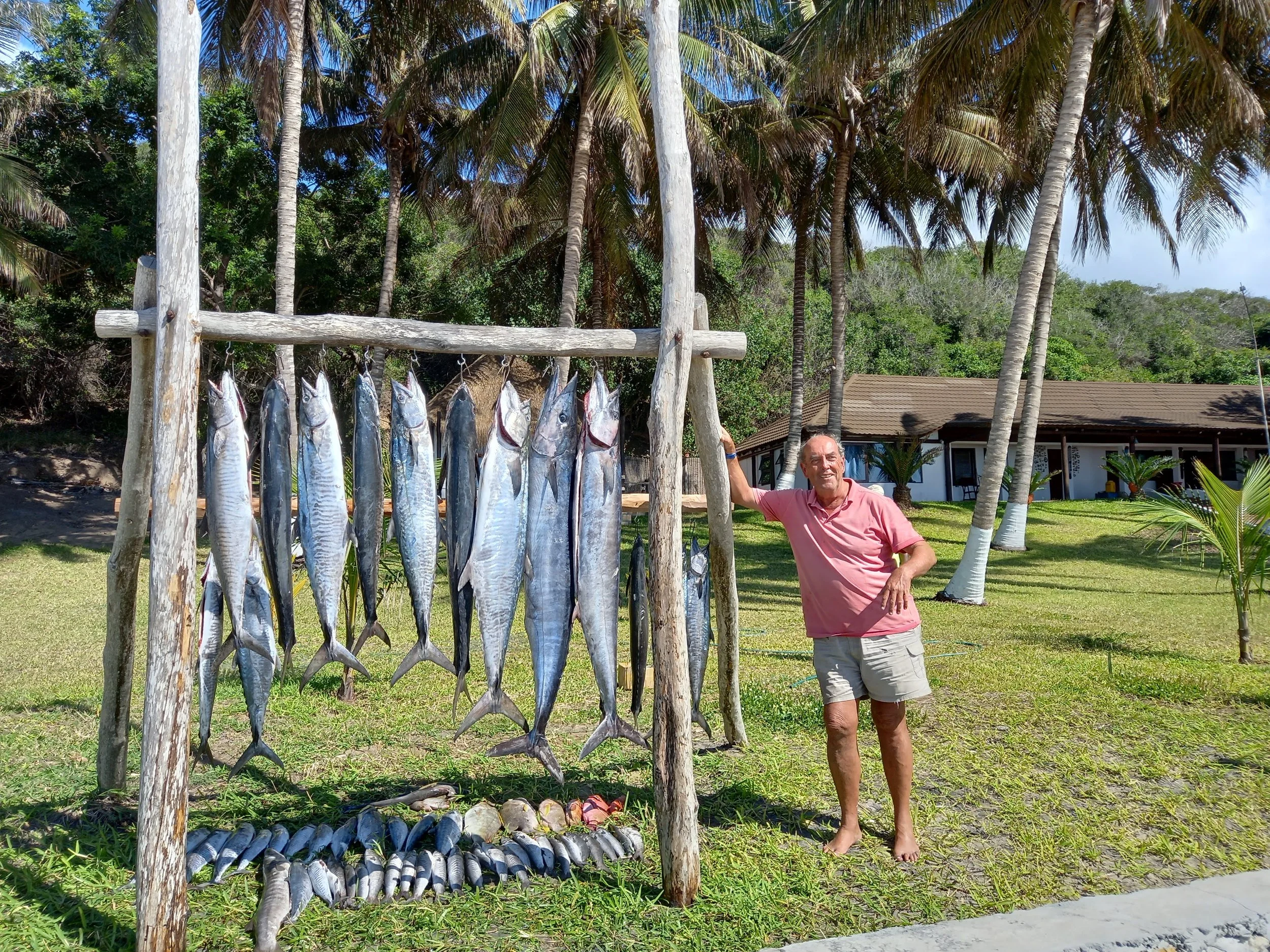 A man standing barefoot on grass, smiling, with a hanging display of fish behind him, surrounded by palm trees and a house in the background.