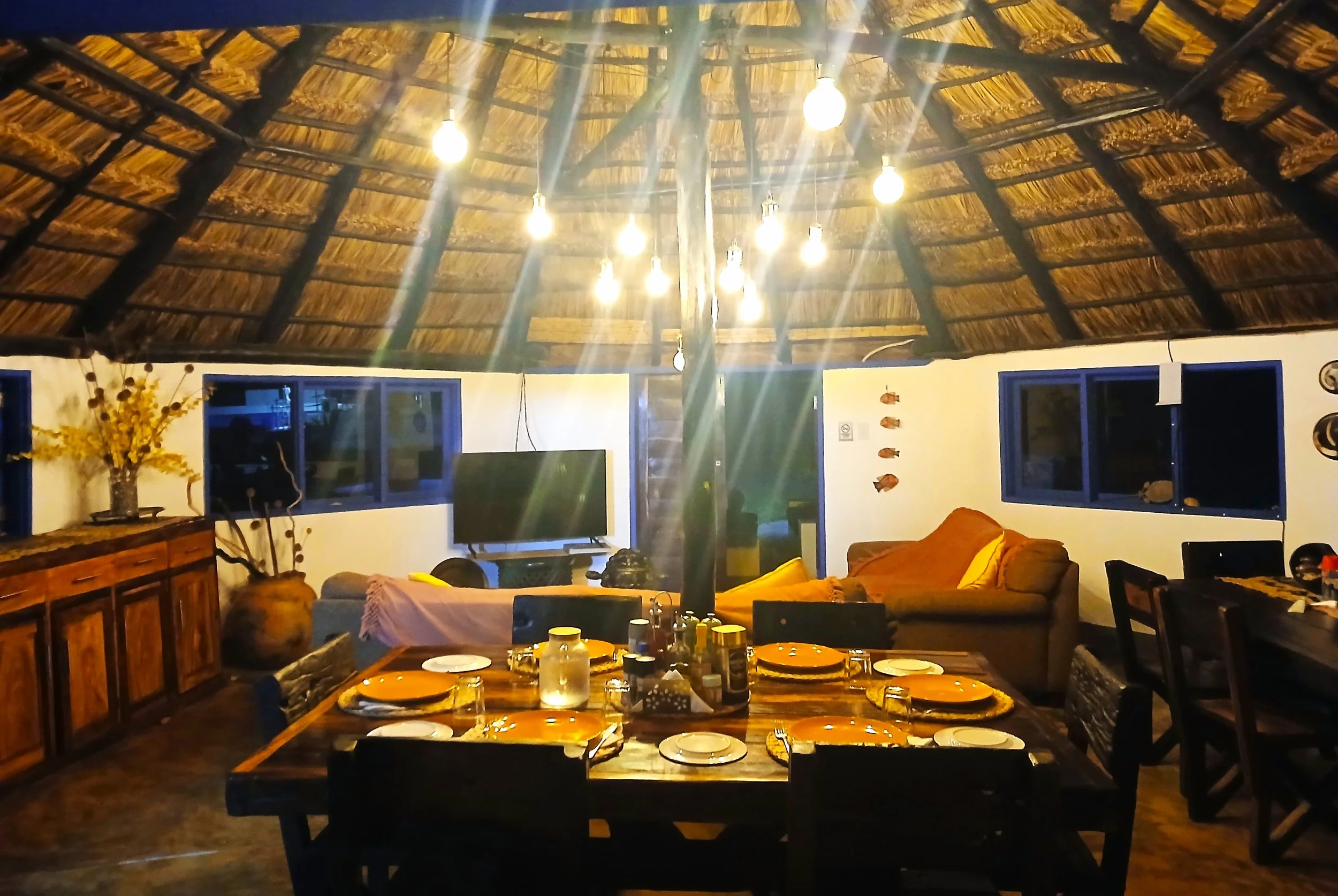 Inside a rustic living room with a thatched ceiling, hanging light bulbs, a wooden dining table set with plates and cups, a gray sofa with yellow and pink cushions, a television, and decorative fish on the wall.