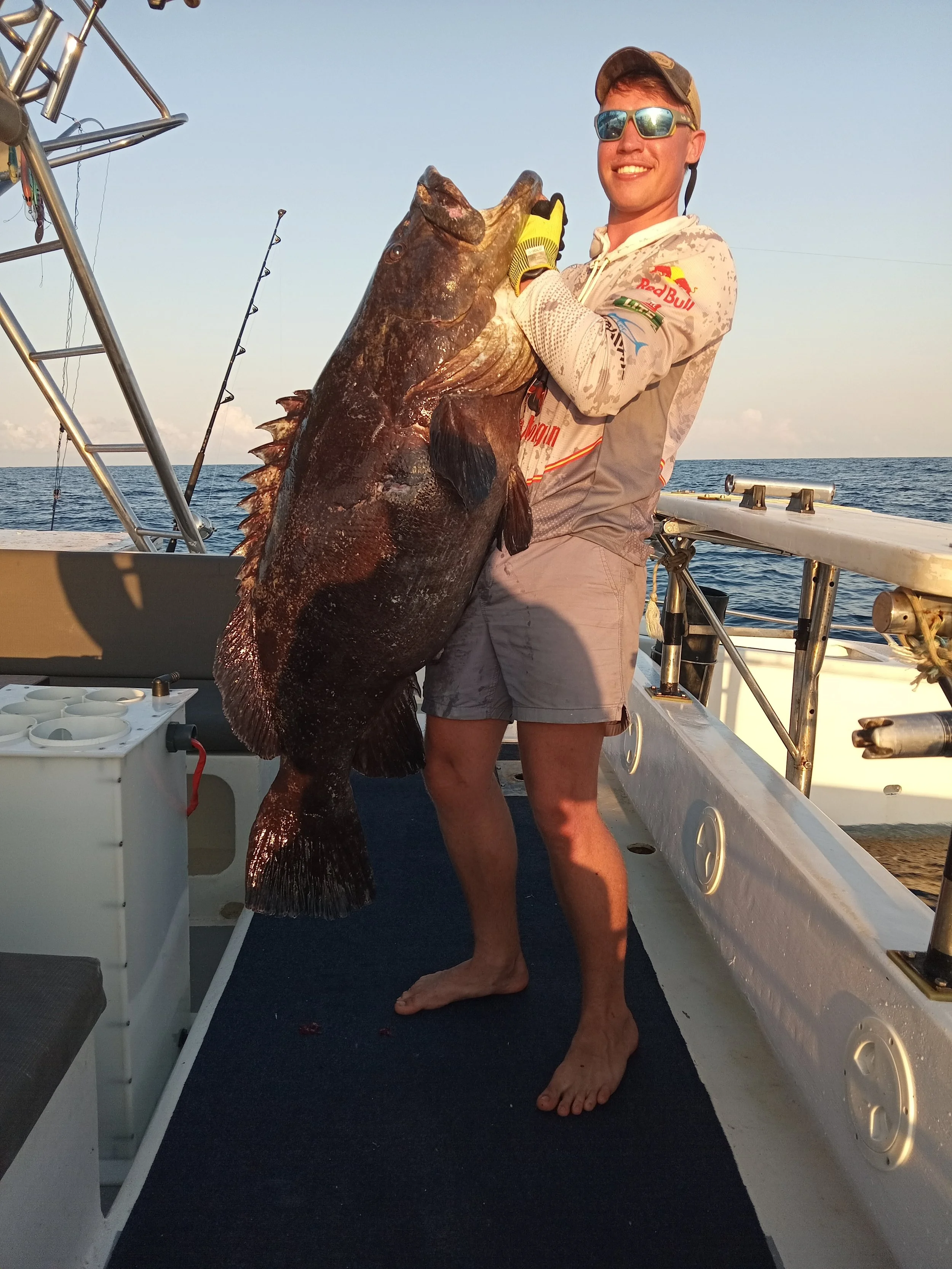 Man holding a large fish on a boat at sea, wearing sunglasses, a hoodie with sports logos, and yellow gloves, smiling at the camera.