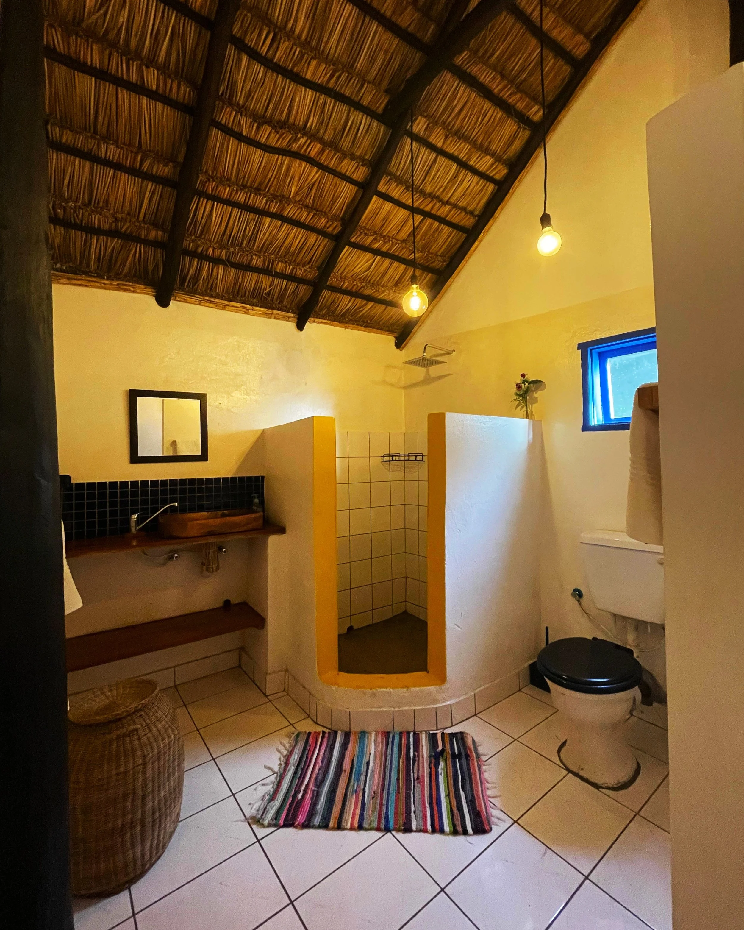 Interior of a room with a thatched roof ceiling, yellow walls, and a small window with blue trim. The room contains a striped rug on tiled floor, a wicker basket, a yellow partition, a small sink, a mirror, a flower vase, and a black stool.