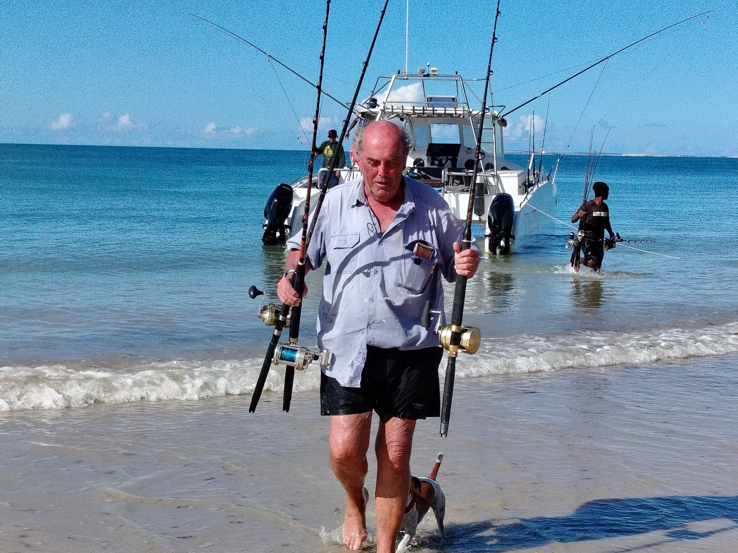 A man holding fishing rods walking out of the sea onto the beach with a boat anchored behind him.