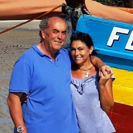 A man and woman smiling and standing close together near a colorful boat at the beach.