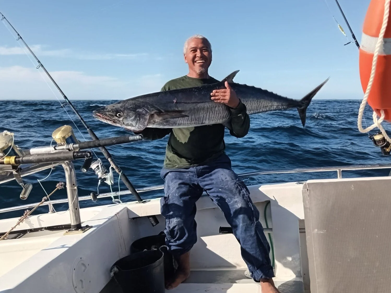 A man holding a large fish on a boat in the ocean, smiling, with fishing rods nearby and a bright blue sky.