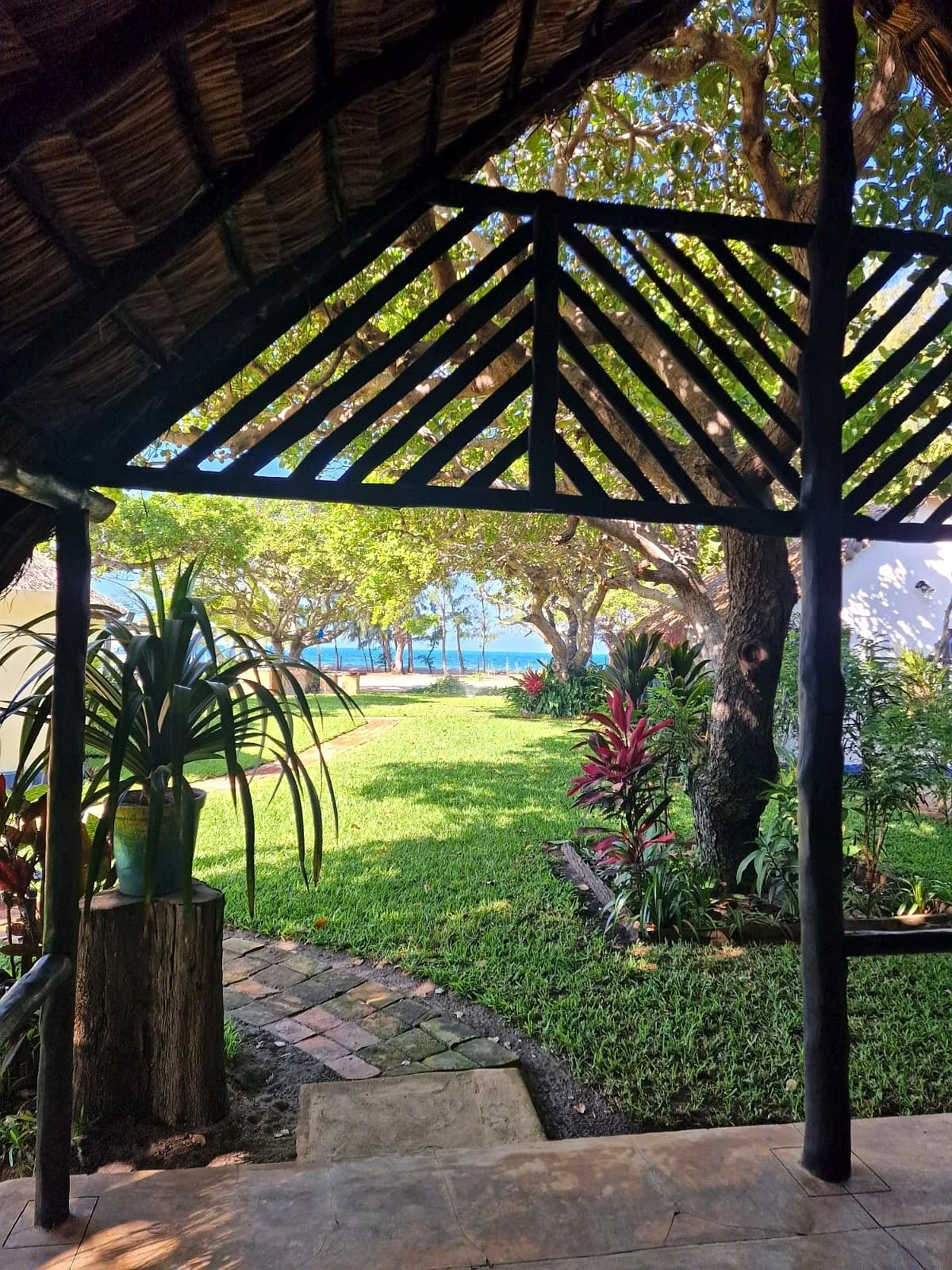 View from a shaded patio with thatched roof, looking out into a lush garden with grass, trees, and tropical plants, and the ocean in the background under a partly cloudy sky.