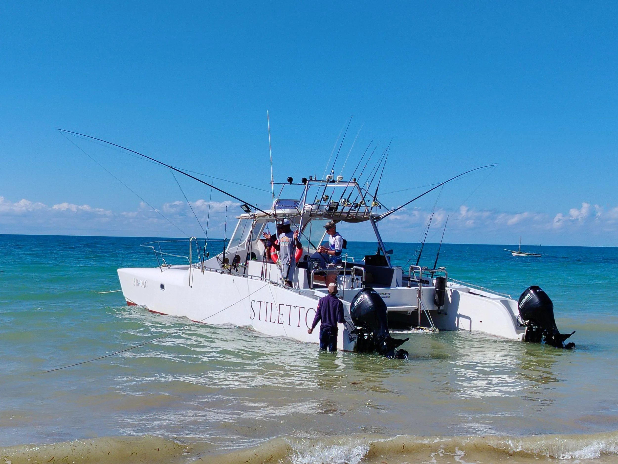 People boarding a white boat named 'STILETTO' in clear shallow water with multiple fishing rods attached, under a bright blue sky.