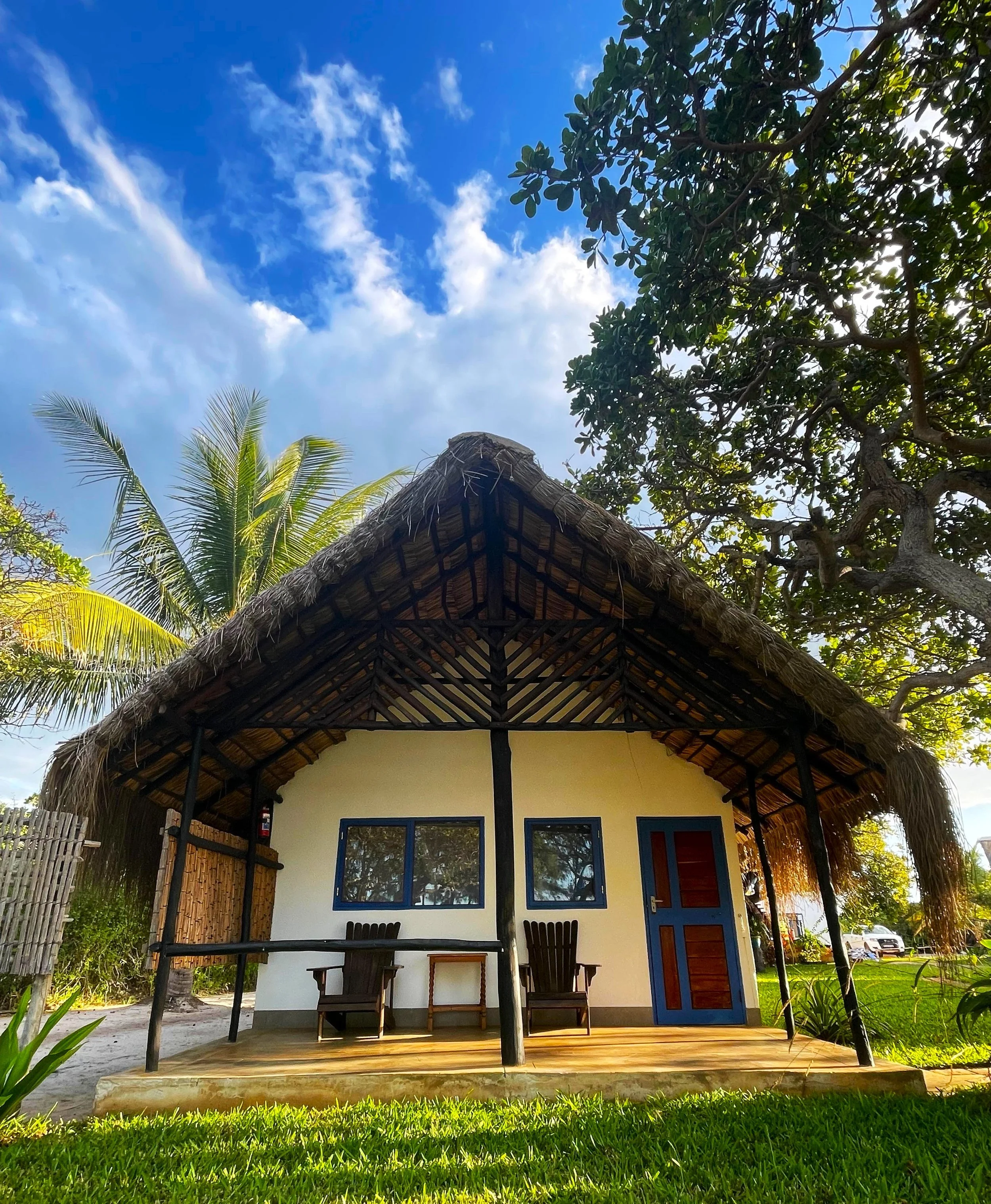 A small tropical house with a thatched roof, blue window frames, and a blue door, surrounded by greenery, palm trees, and clear blue sky with some clouds.